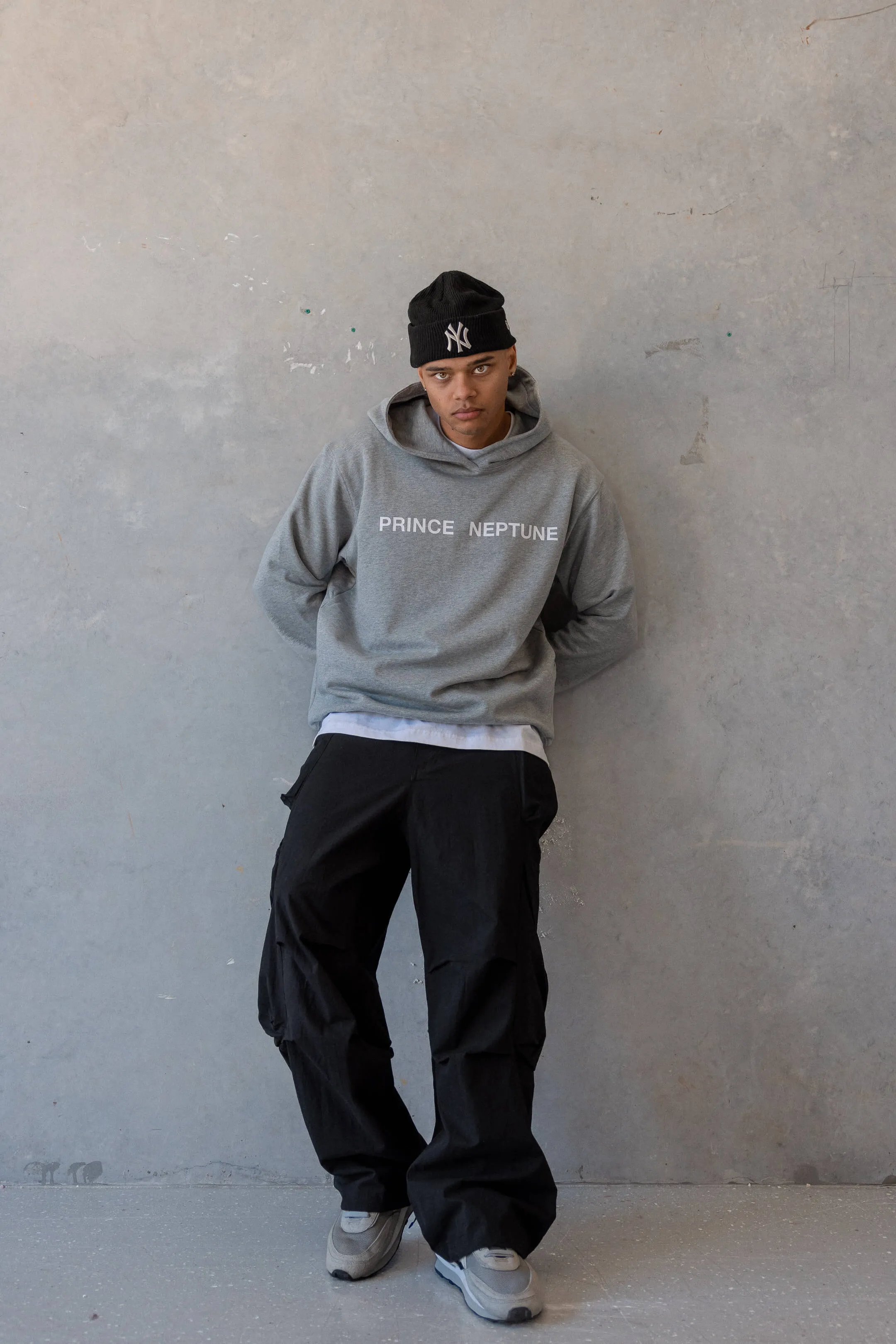 Model leaning against wall in grey Prince Neptune hoodie — Urban streetwear portrait of a model wearing a grey “Prince Neptune” hoodie and black pants with a black beanie, from the 2023 Collection One editorial | EDG Studios