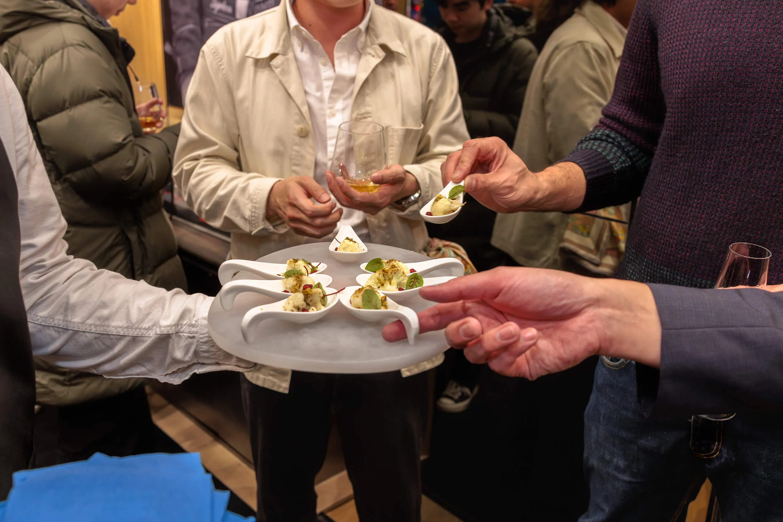 Close-up of a catering tray with modern white spoons serving a bite-sized canapé as guests reach for the hors d’oeuvres with drinks in hand | EDG Studios