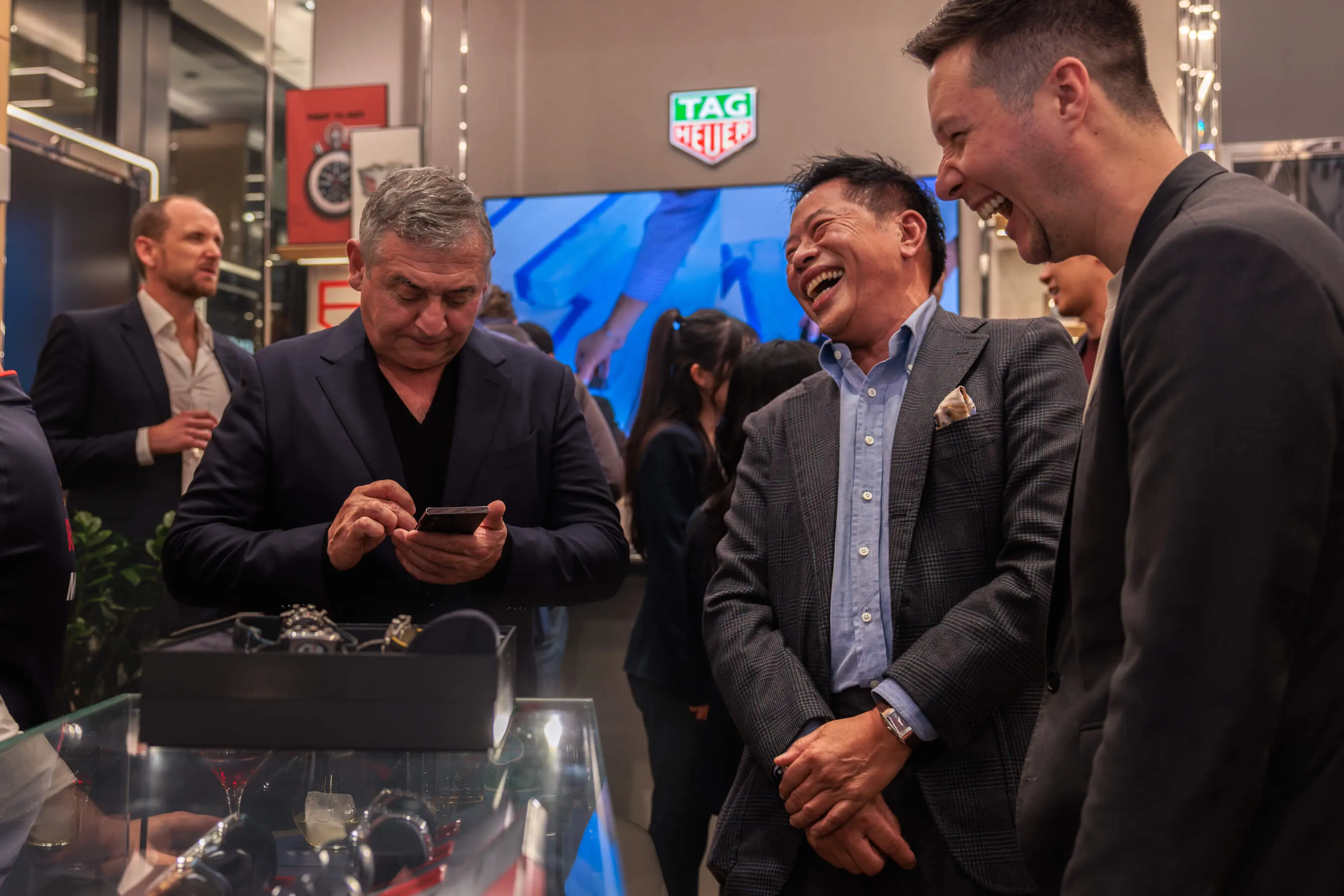 A group of well-dressed men share a laugh around a glass display of watches during the evening event, with a TAG Heuer logo illuminated in the background | EDG Studios