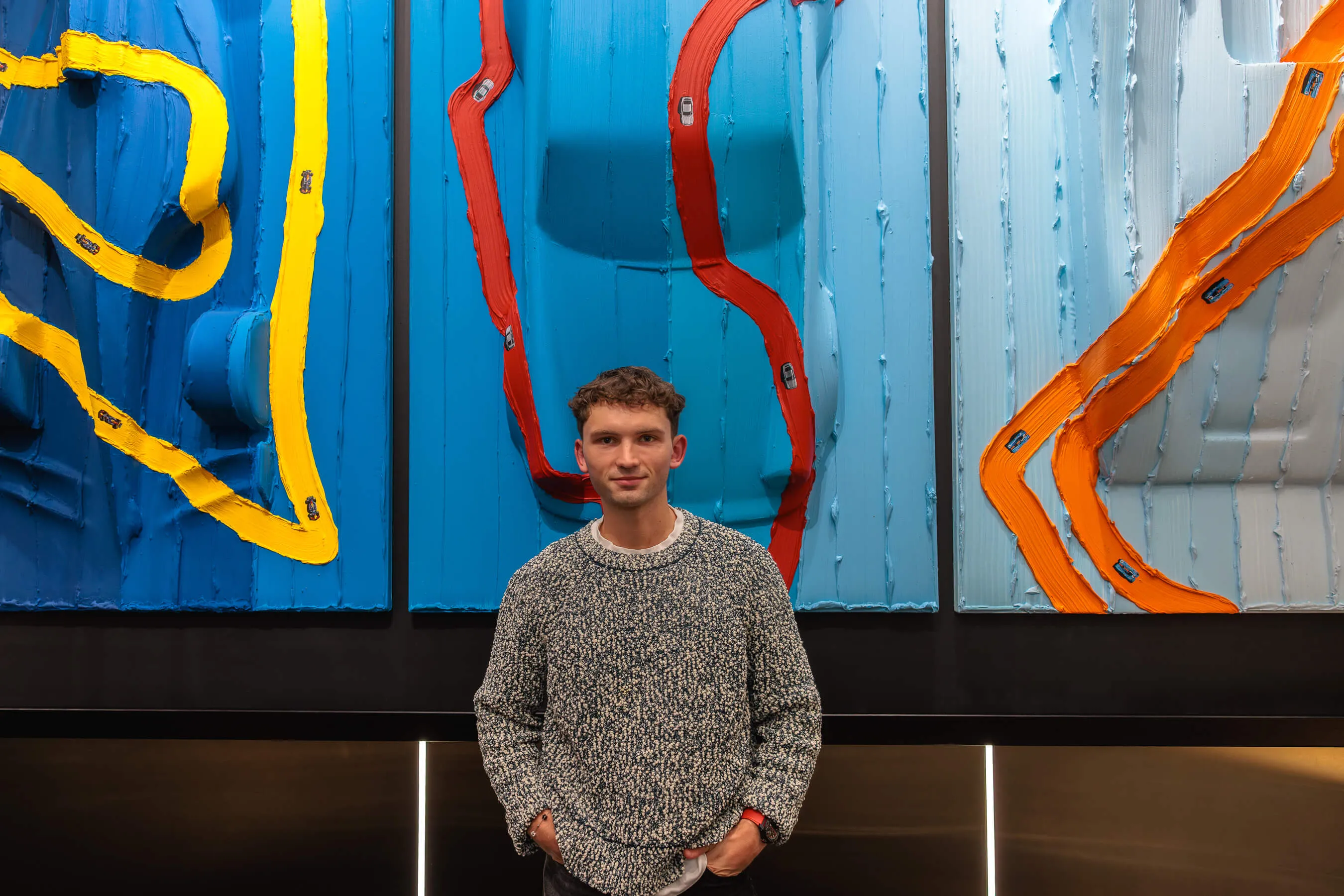 Artist Werner Bronkhorst stands in front of his colourful triptych artwork featuring toy cars and winding painted roads in yellow, red, and orange on blue-toned panels | EDG Studios