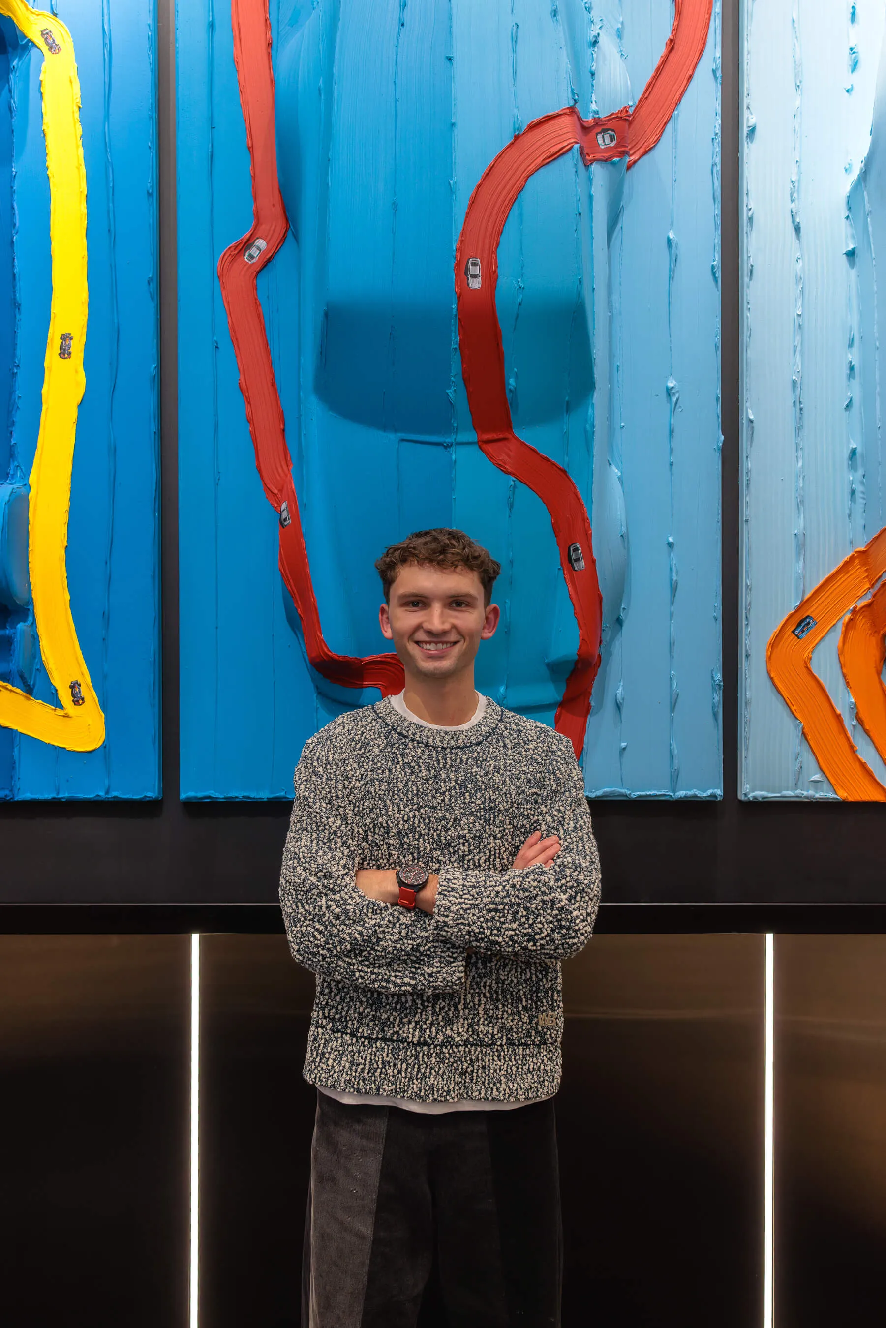 Werner Bronkhorst smiles in front of his artwork, arms crossed, wearing a red-banded TAG Heuer timepiece against the textured panels | EDG Studios