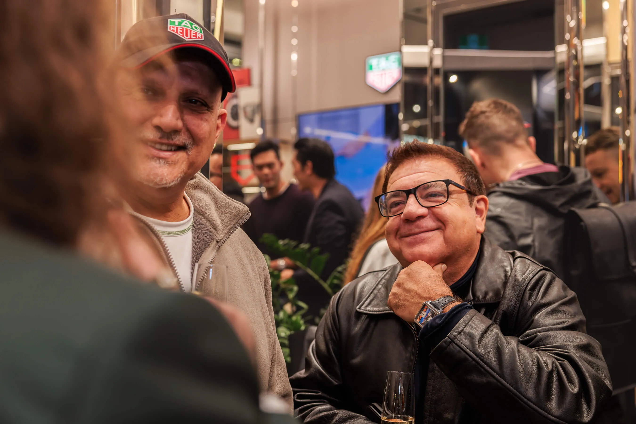 Two guests chat and smile at the launch event, one wearing a TAG Heuer cap and the other in glasses and a leather jacket, showing off a colorful TAG Heuer watch | EDG Studios