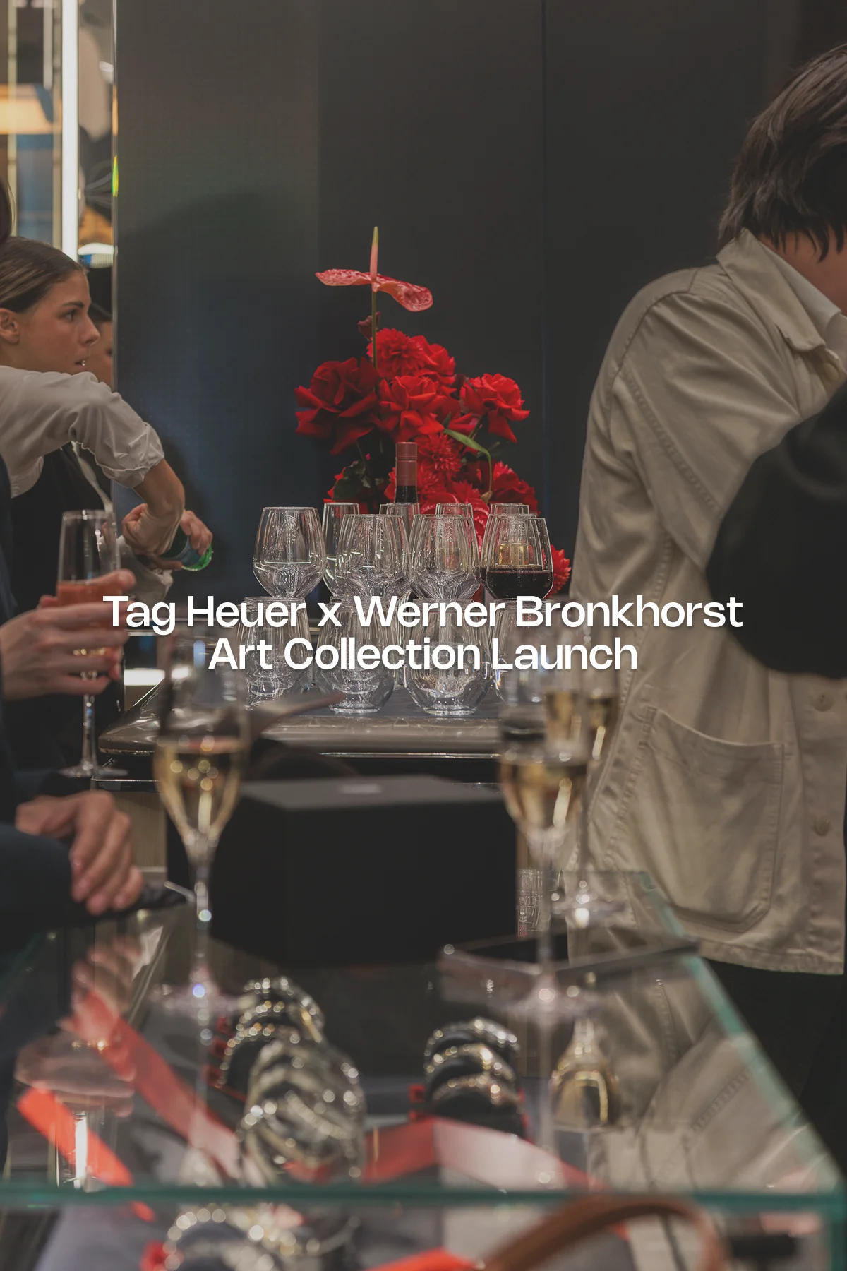 Glasses of champagne and wine are arranged beside a bold floral arrangement of red roses and anthuriums, with the overlay text “TAG HEUER × WERNER BRONKHORST” marking the luxury collaboration event; reflections of high-end watches and guests surround the bar setup.