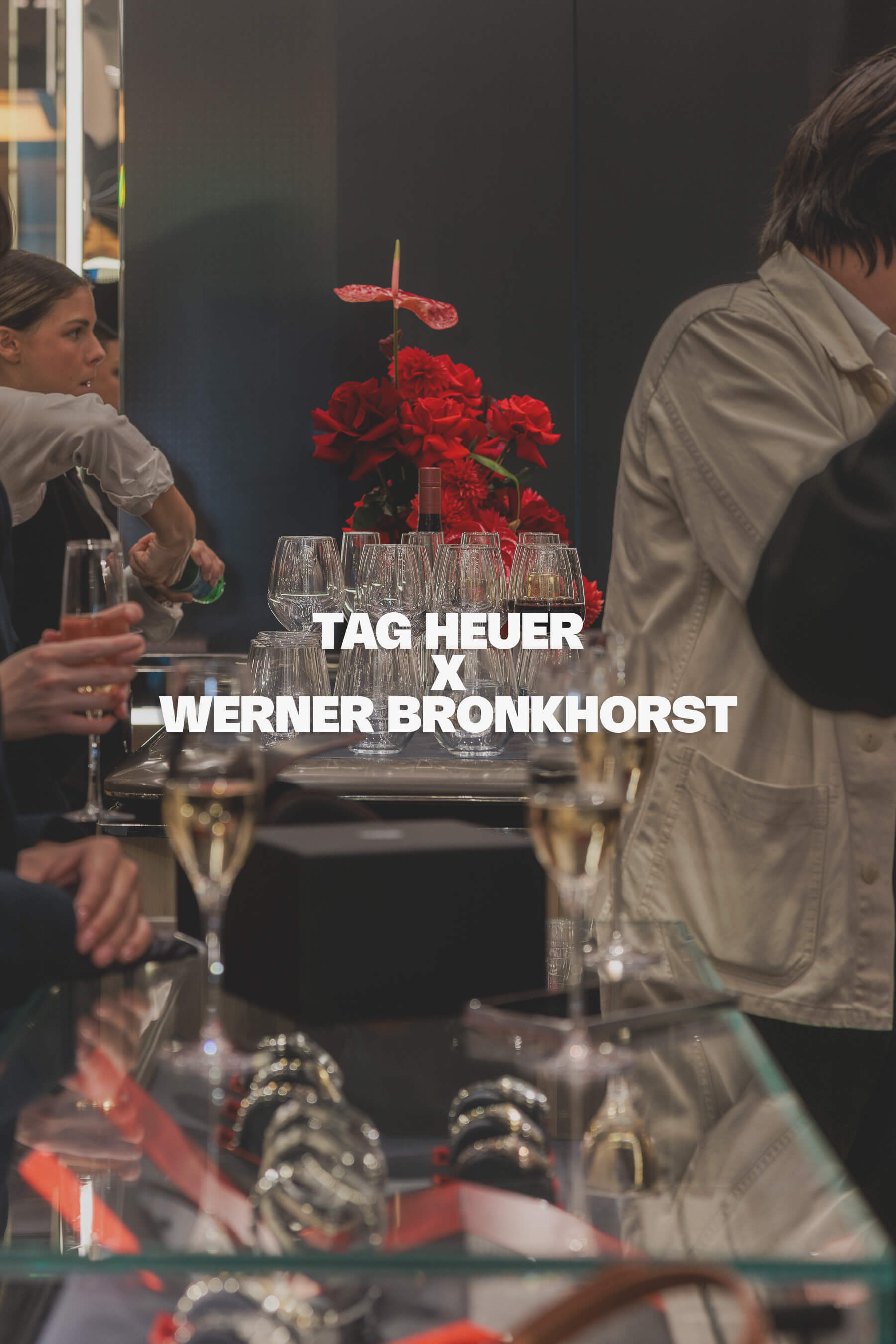 Glasses of champagne and wine are arranged beside a bold floral arrangement of red roses and anthuriums, with the overlay text “TAG HEUER × WERNER BRONKHORST” marking the luxury collaboration event; reflections of high-end watches and guests surround the bar setup.