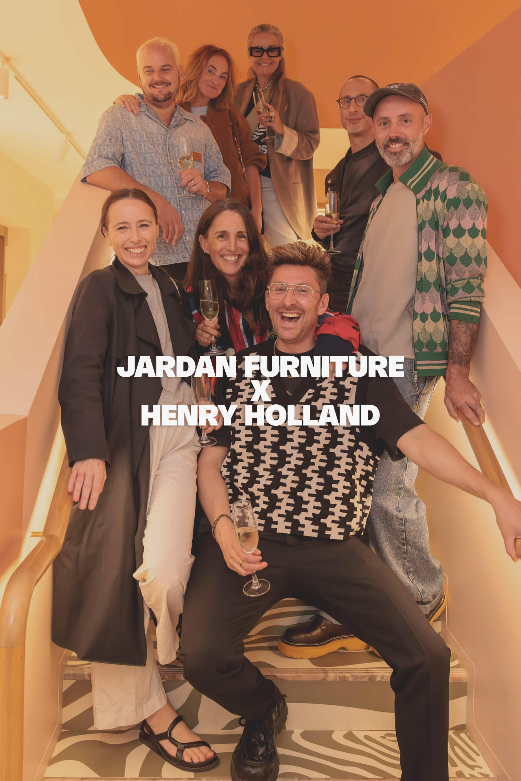 A group of eight stylish guests, including Henry Holland, pose on a bold graphic staircase at the Jardan showroom, smiling and holding champagne flutes during the 2024 Jardan x Henry Holland collaboration event.