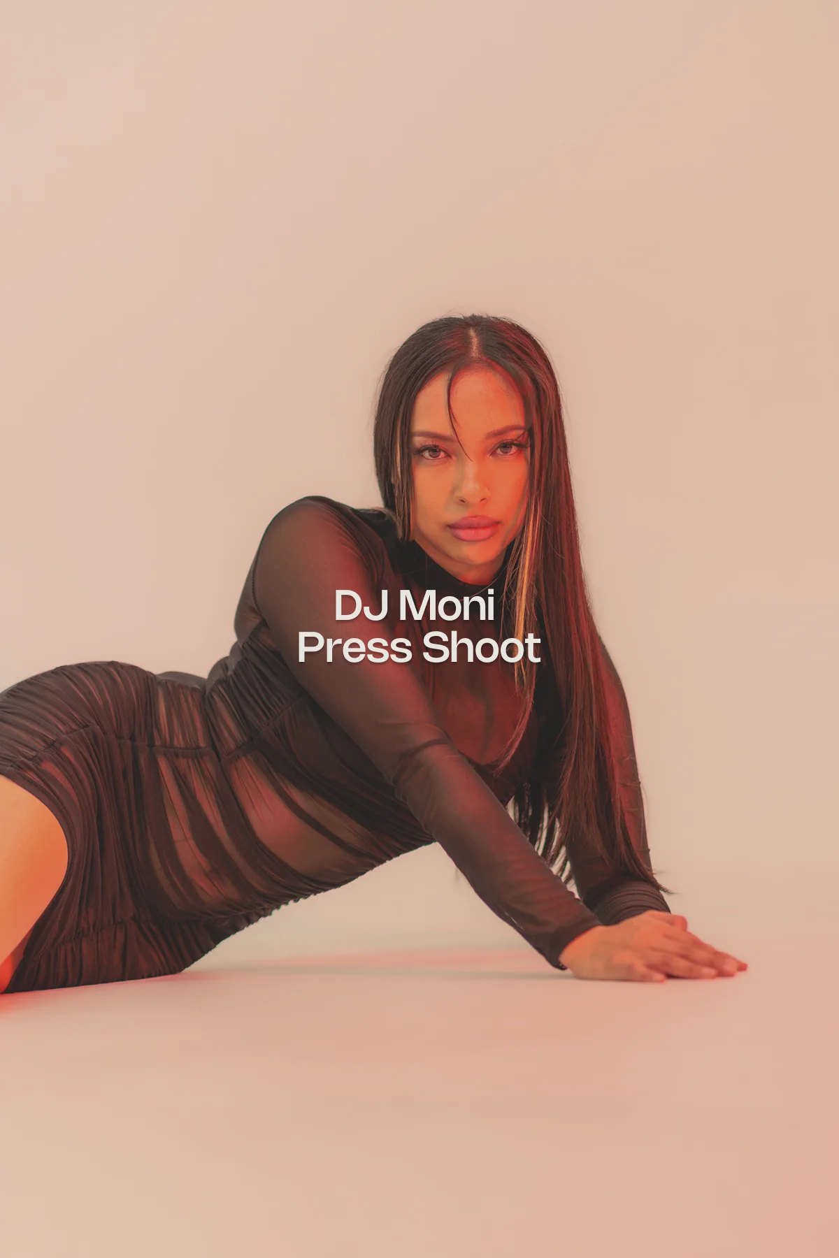 DJ Moni poses against a softly lit peach-pink background wearing a cropped white shirt, black tie, and pleated mini skirt—captured in a bold, stylised editorial portrait.