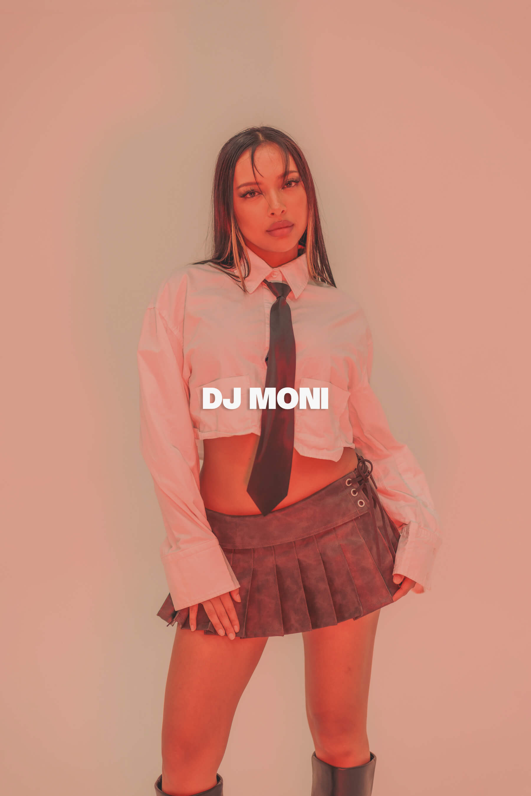 DJ Moni poses against a softly lit peach-pink background wearing a cropped white shirt, black tie, and pleated mini skirt—captured in a bold, stylised editorial portrait.
