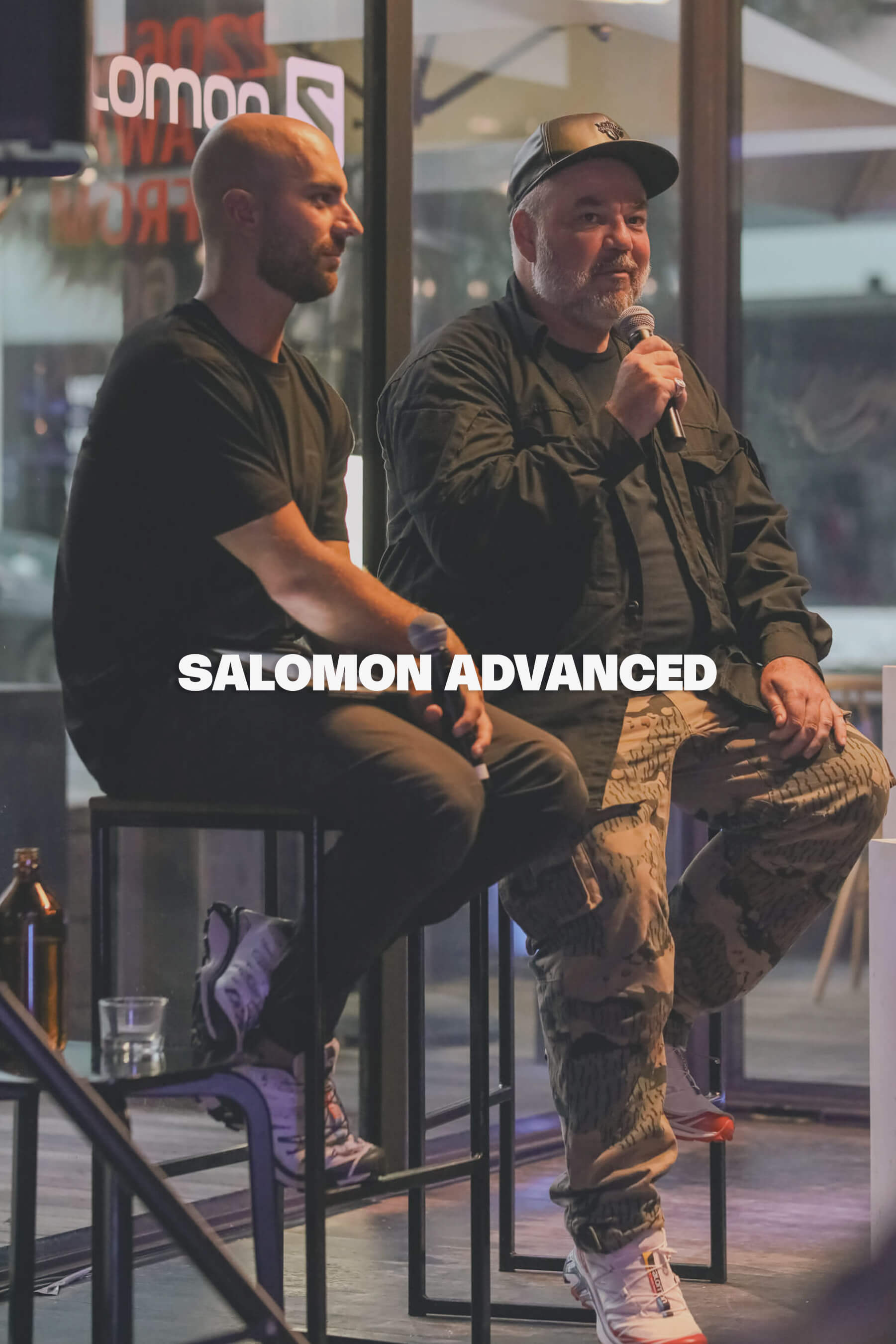 Woody from Sneaker Freaker speaks into a microphone during the Salomon Advanced season launch event, seated beside a fellow panelist on stage with sneakers on display and branding reflected in the venue’s glass backdrop.