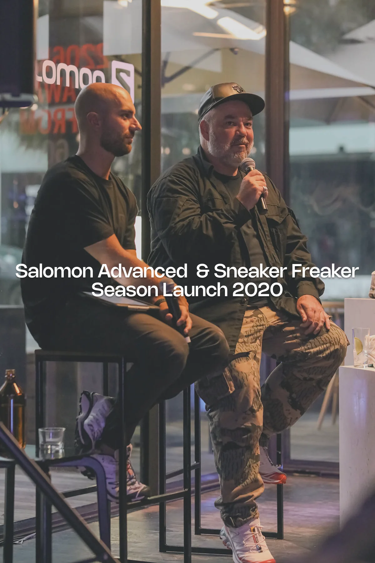 Woody from Sneaker Freaker speaks into a microphone during the Salomon Advanced season launch event, seated beside a fellow panelist on stage with sneakers on display and branding reflected in the venue’s glass backdrop.