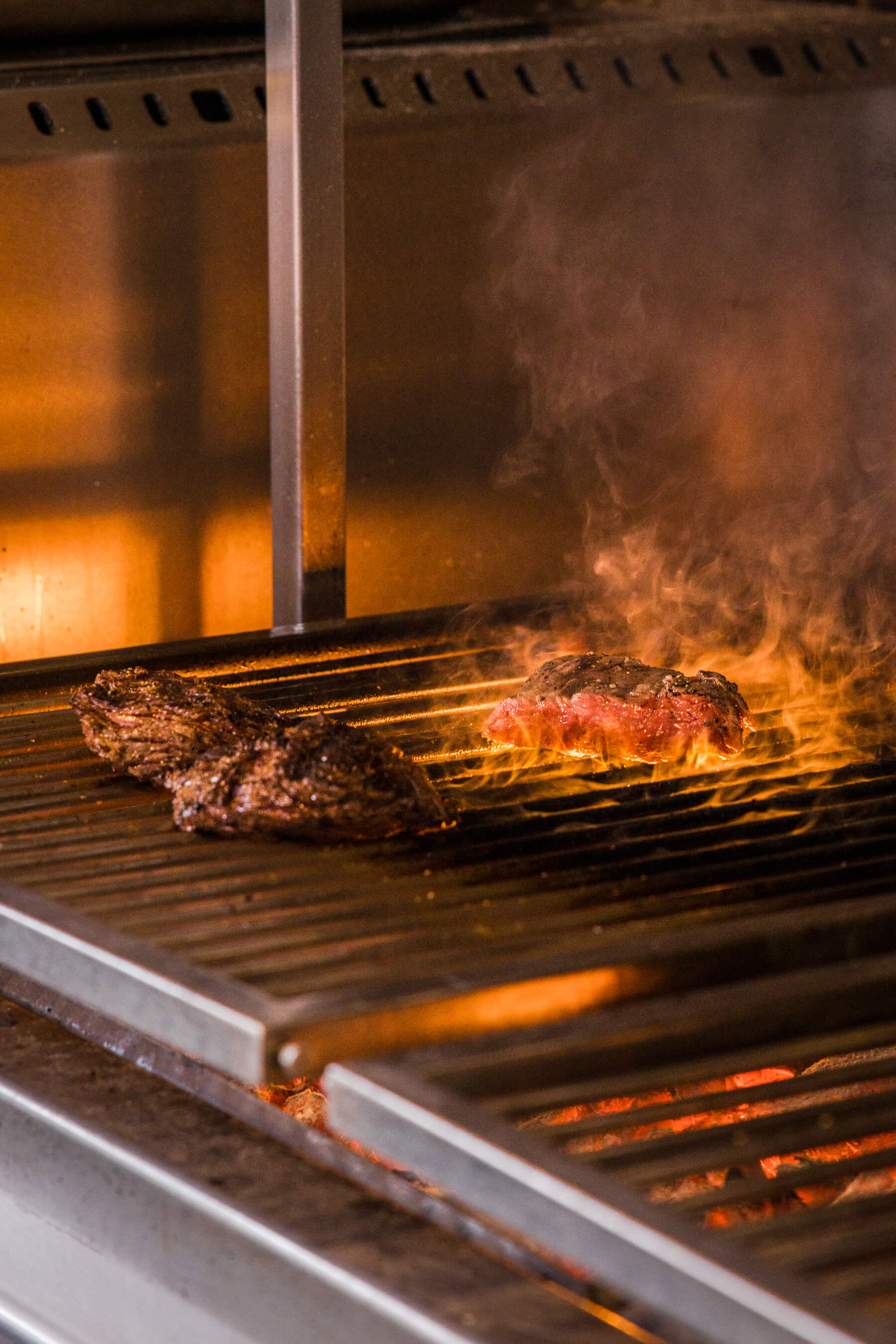 Charred steaks cooking over glowing coals and open flames at Le Foote, dramatic lighting capturing the fire's intensity