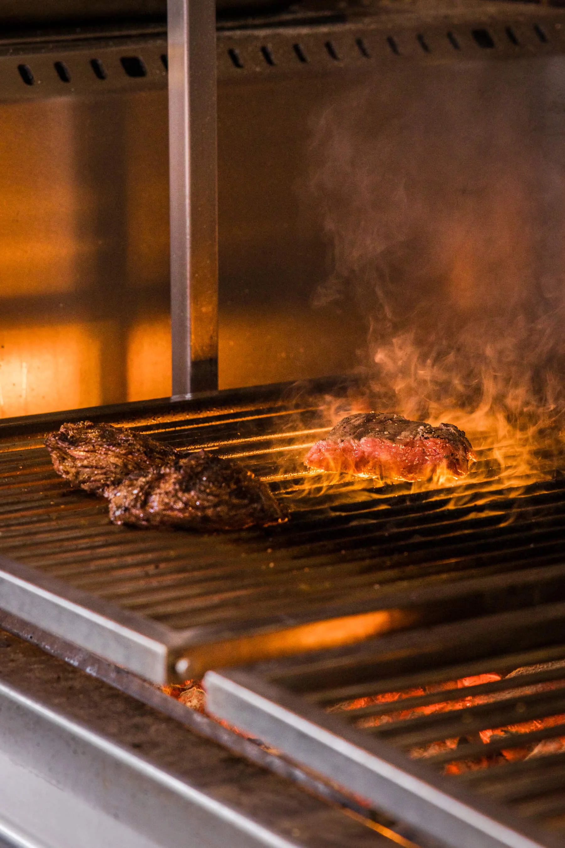 Charred steaks cooking over glowing coals and open flames at Le Foote, dramatic lighting capturing the fire's intensity | EDG Studios