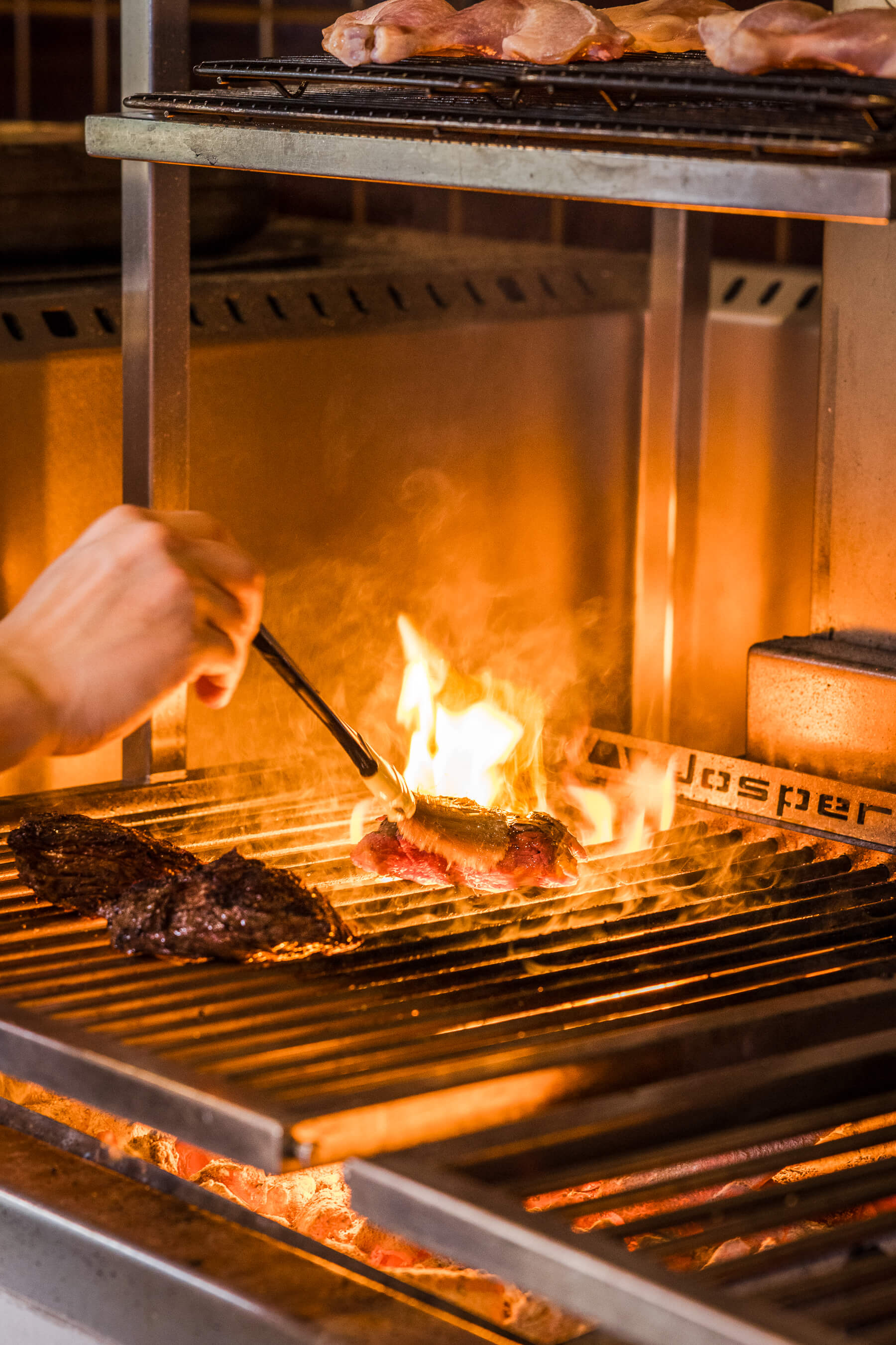 Multiple cuts of meat cooking on the Josper grill at Le Foote, Sydney, with flames rising under the chef’s hand