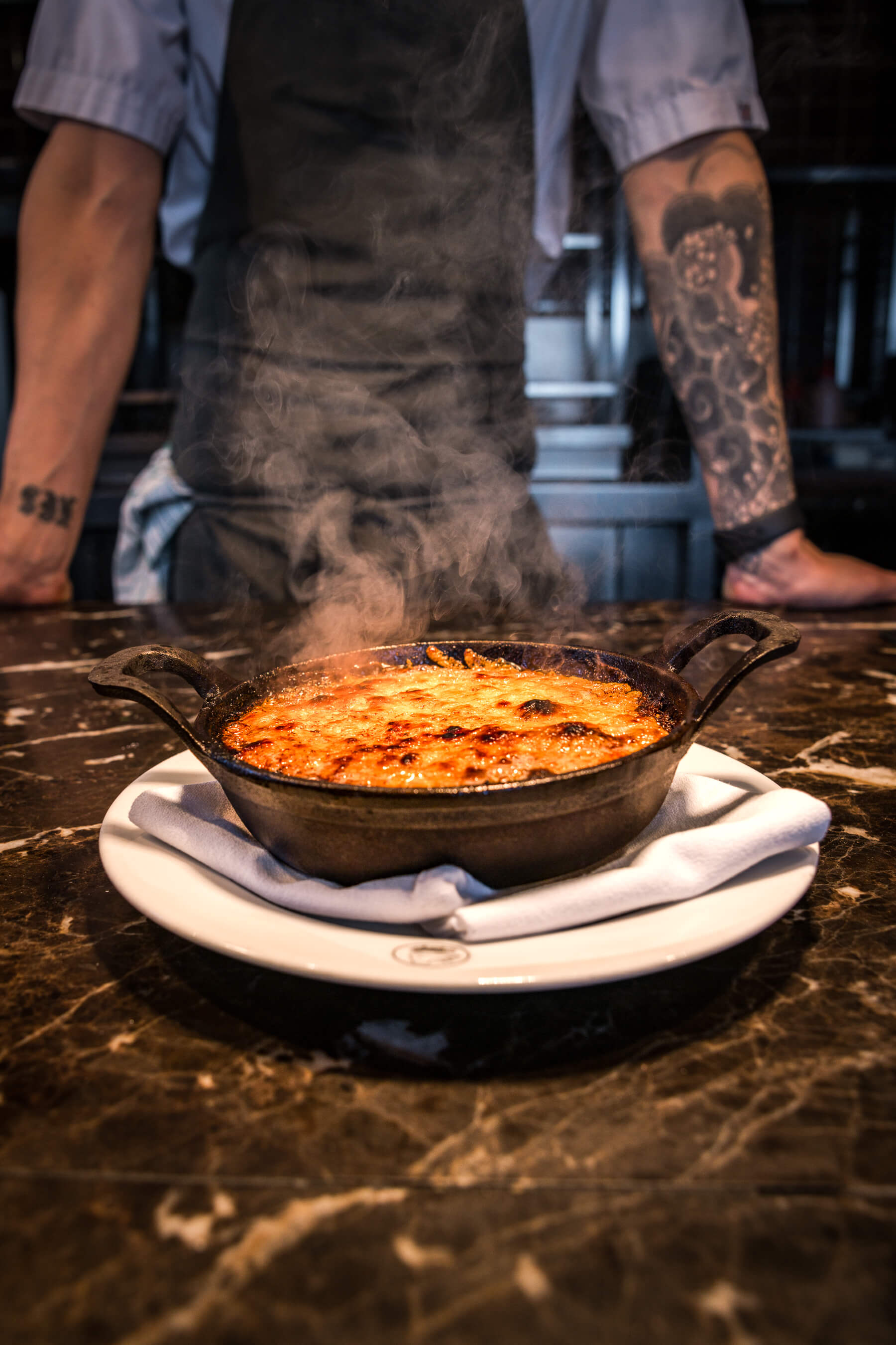 Chef with tattooed arms standing behind a signature steaming dish on the counter at Le Foote, Sydney