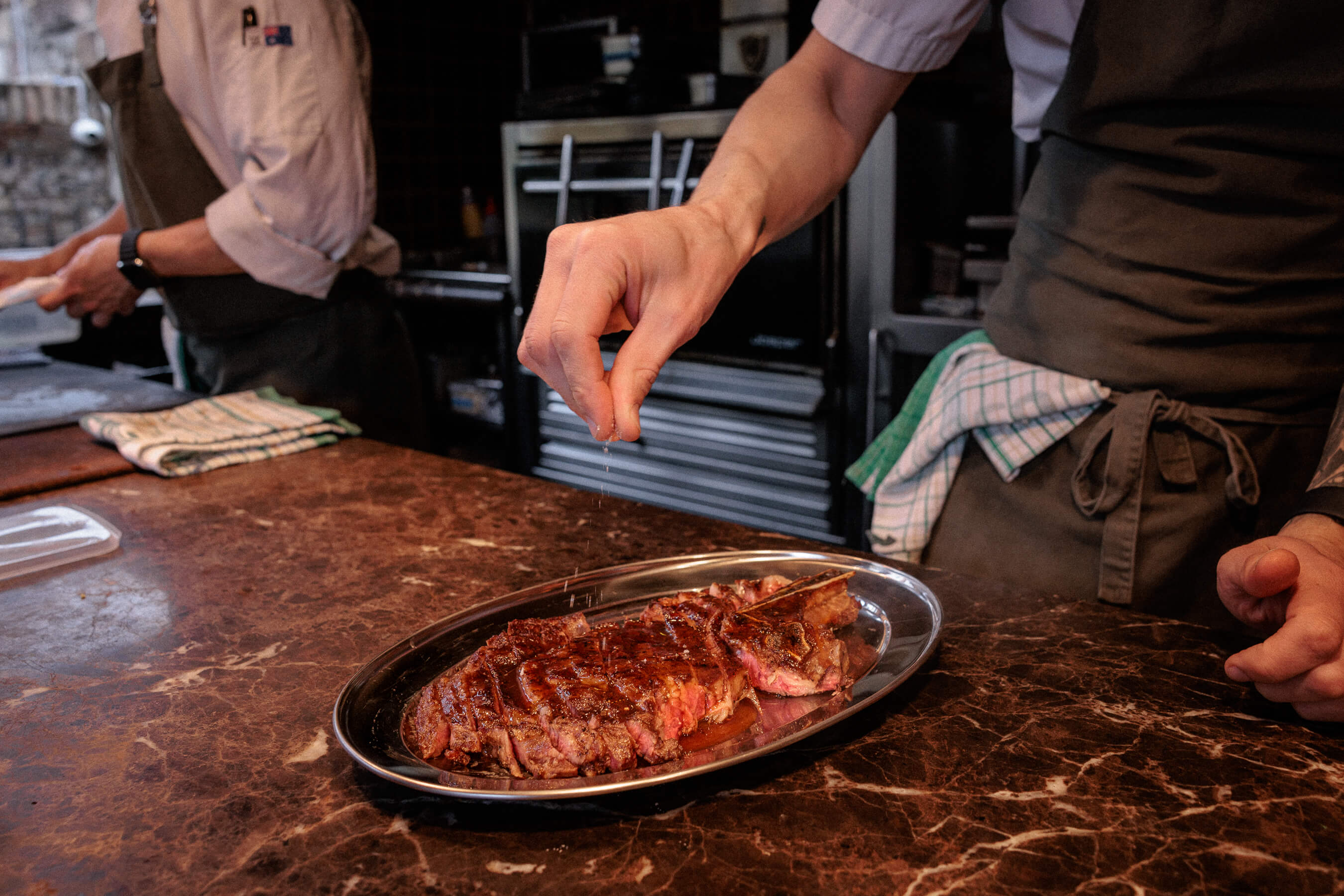 Close-up of chef seasoning a sliced steak with sea salt at Le Foote Sydney, rustic kitchen setting in background