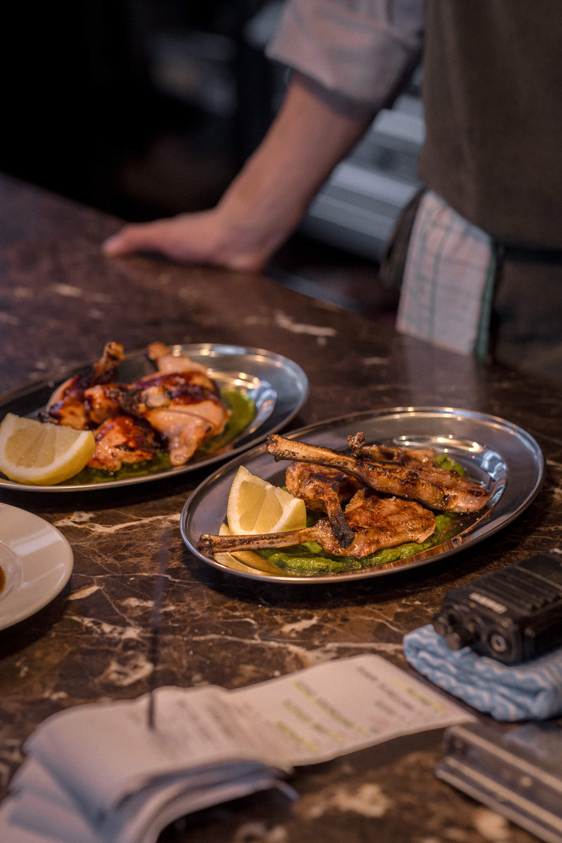 Two plated dishes—grilled lamb and chicken—on marble pass with chef in background at Le Foote, kitchen service setting