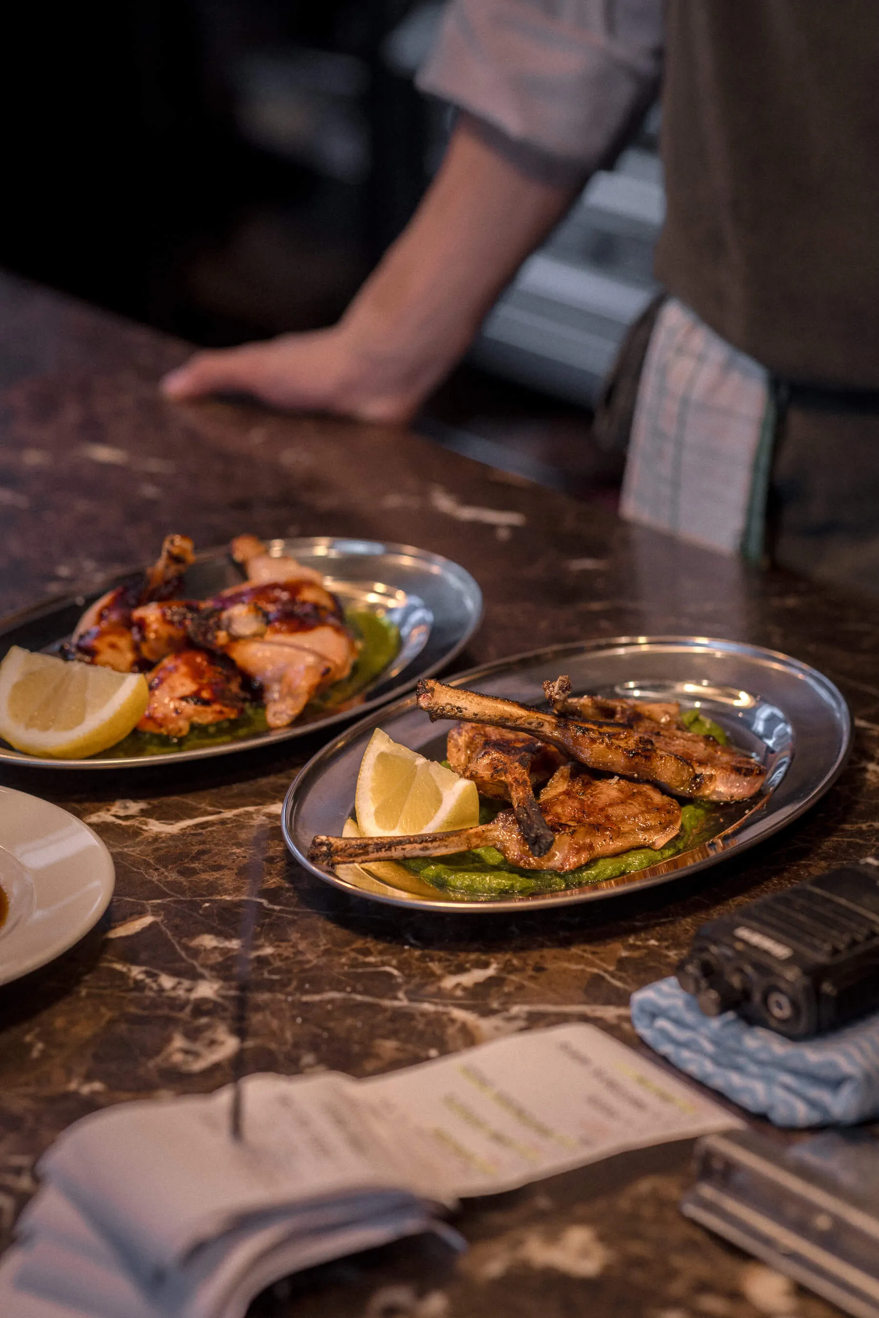 Two plated dishes—grilled lamb and chicken—on marble pass with chef in background at Le Foote, kitchen service setting | EDG Studios
