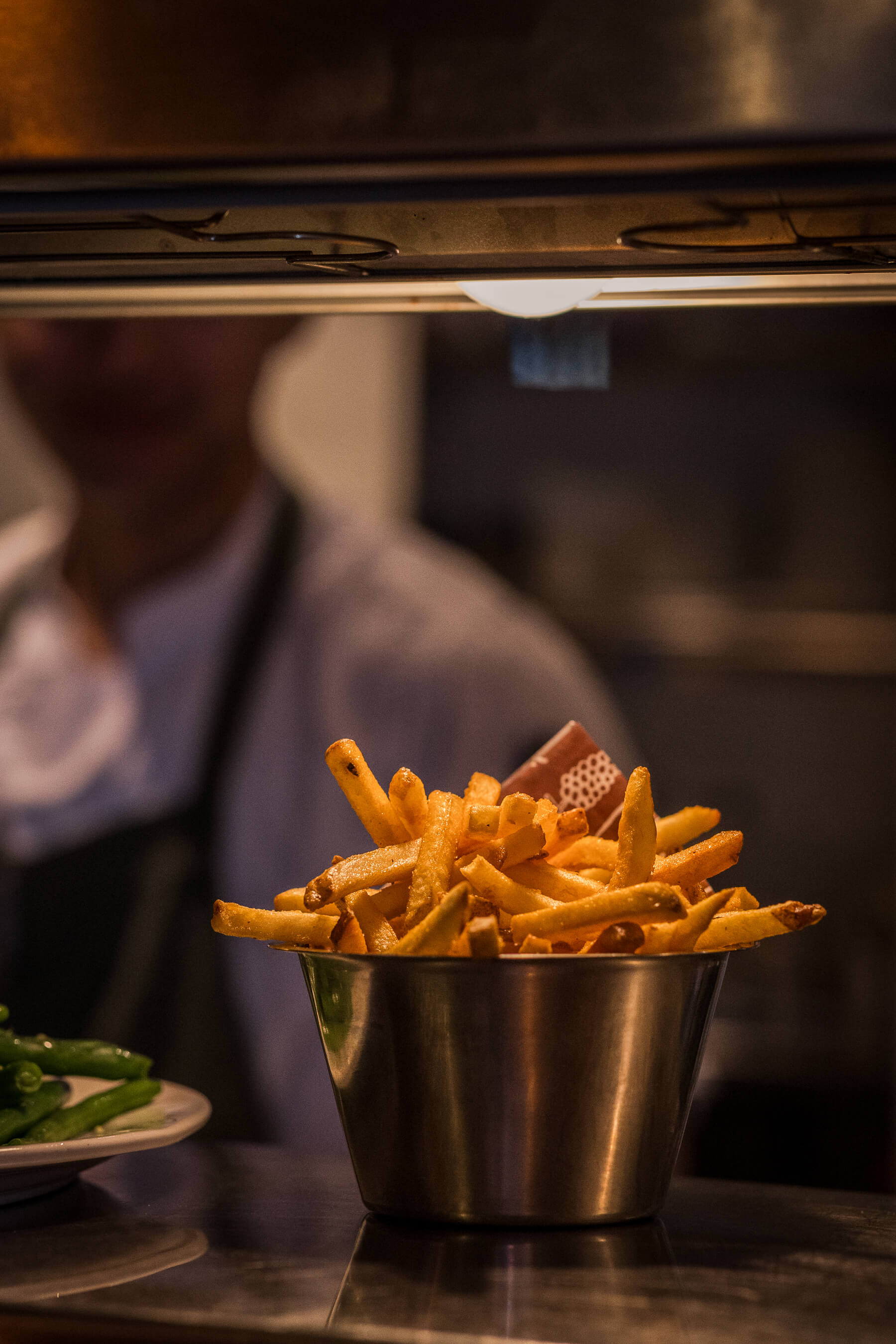 Golden fries served in a stainless steel bucket under kitchen heat lamps at Le Foote Sydney, chef blurred in background