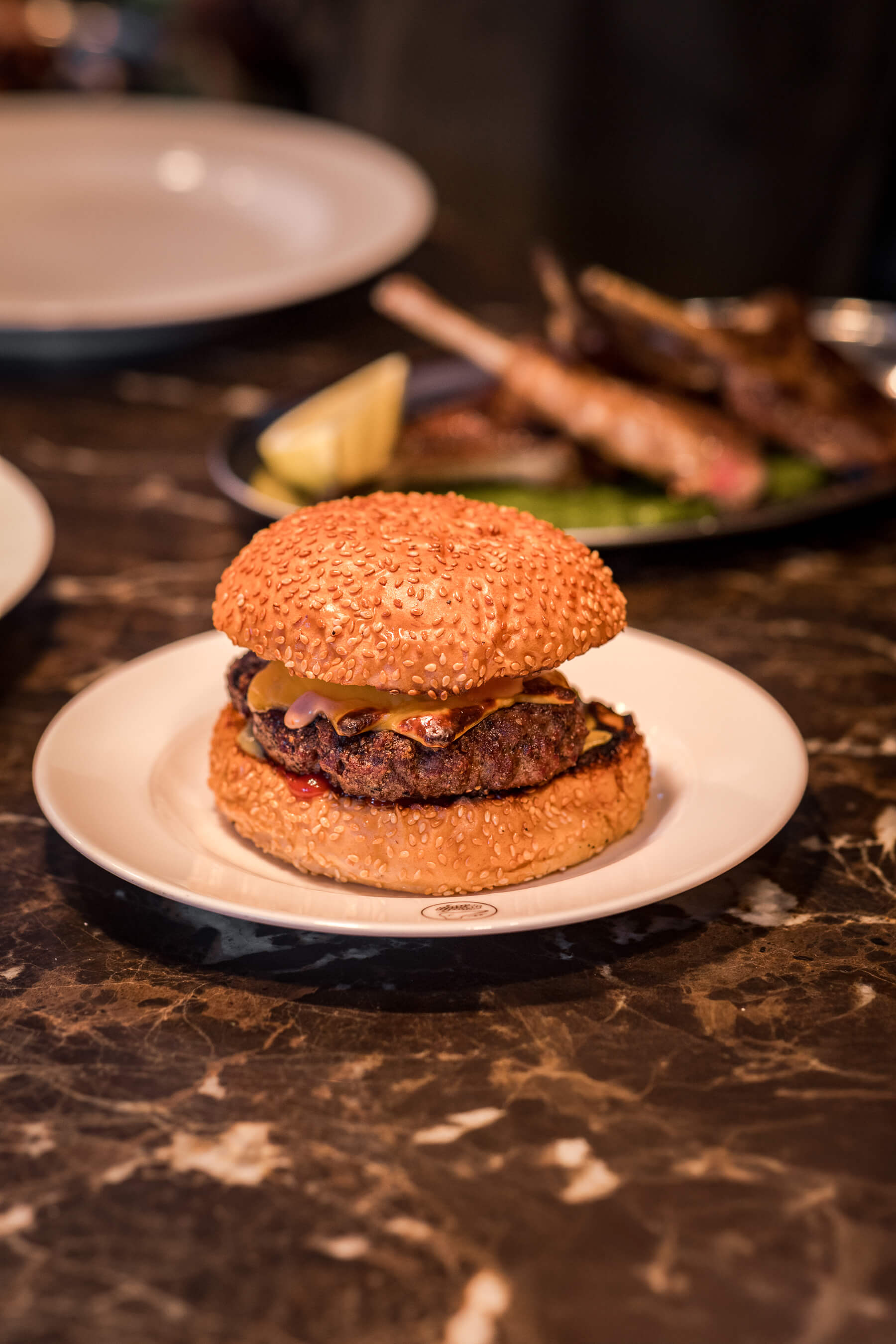 Le Foote’s signature cheeseburger served on sesame seed bun, plated on marble countertop with lamb chops in background
