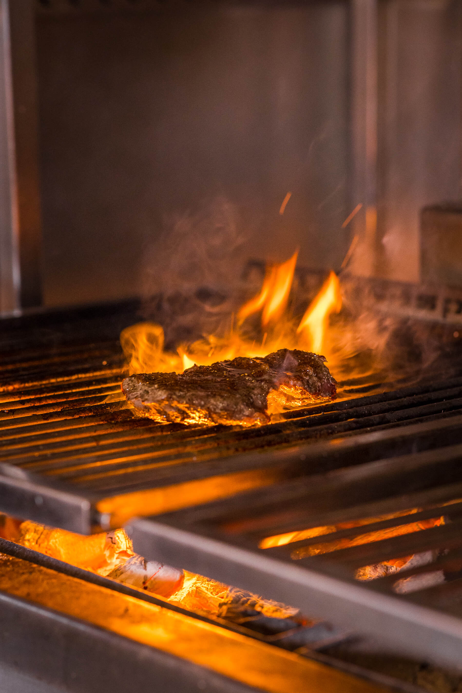 Close-up of ribeye steak cooking over live fire grill with rising smoke at Le Foote’s open kitchen