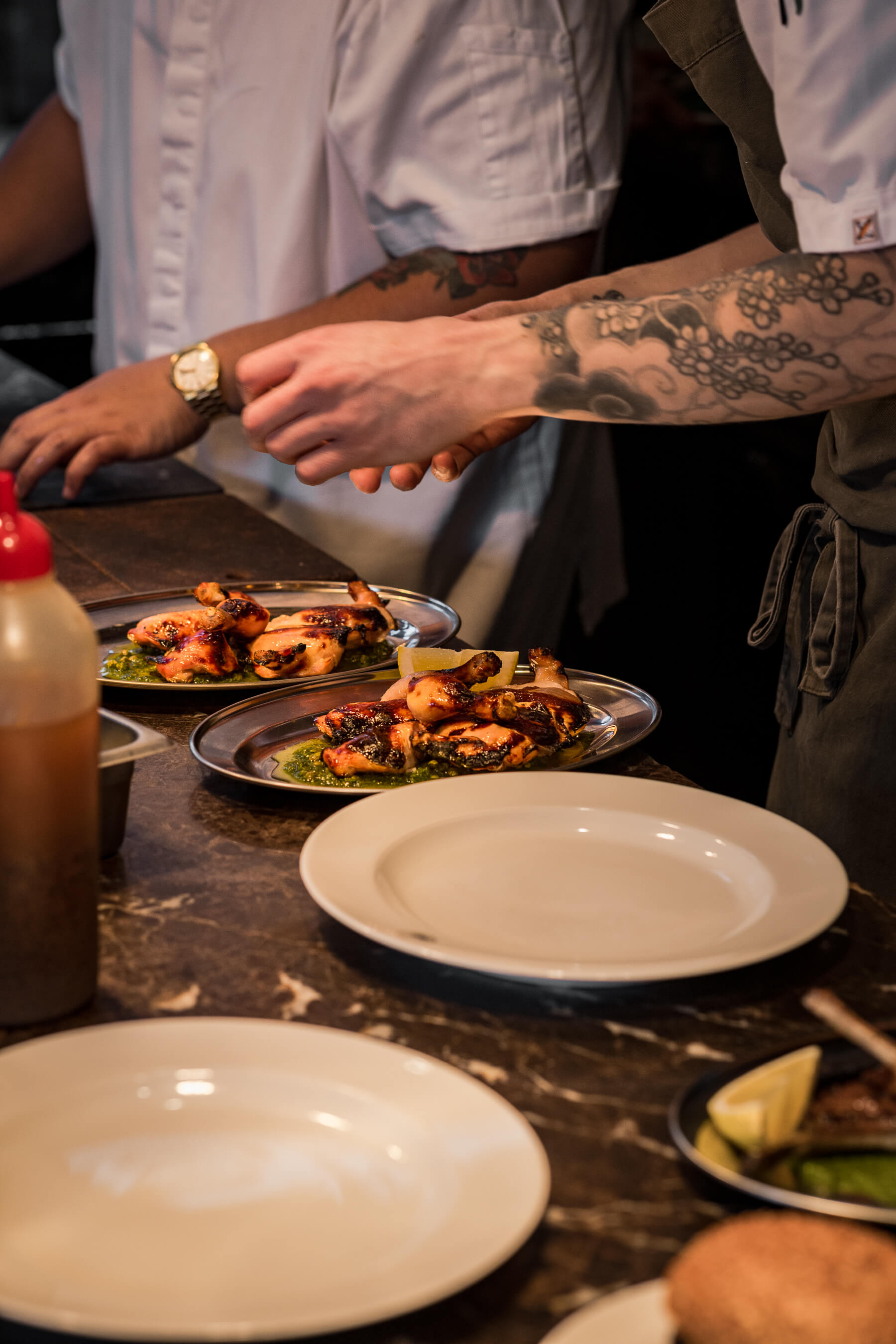 Chef’s tattooed hand plating chargrilled chicken over chimichurri at Le Foote during live kitchen service
