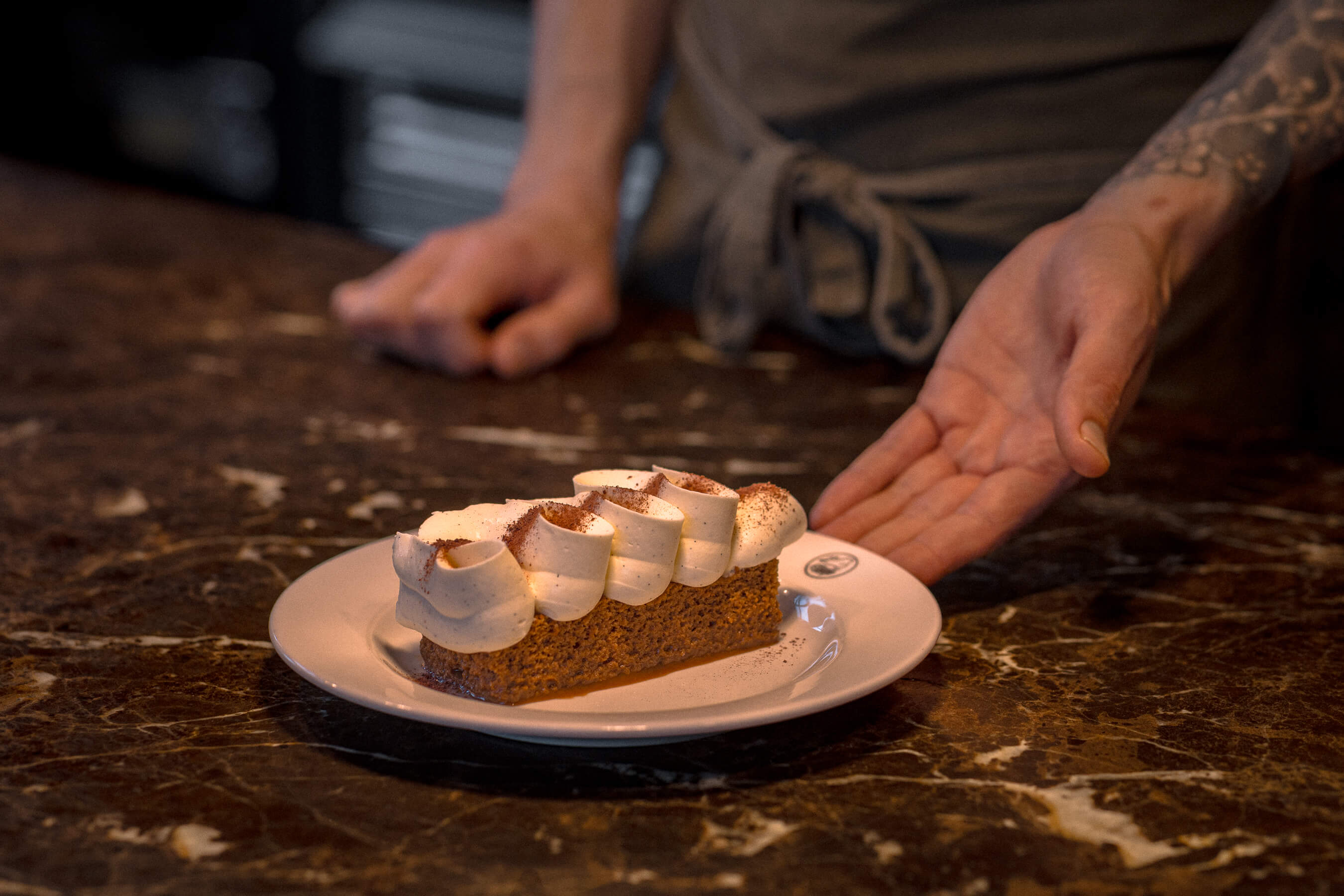 Carrot cake slice topped with rich cream frosting, presented by chef’s tattooed hand at Le Foote on a marble pass