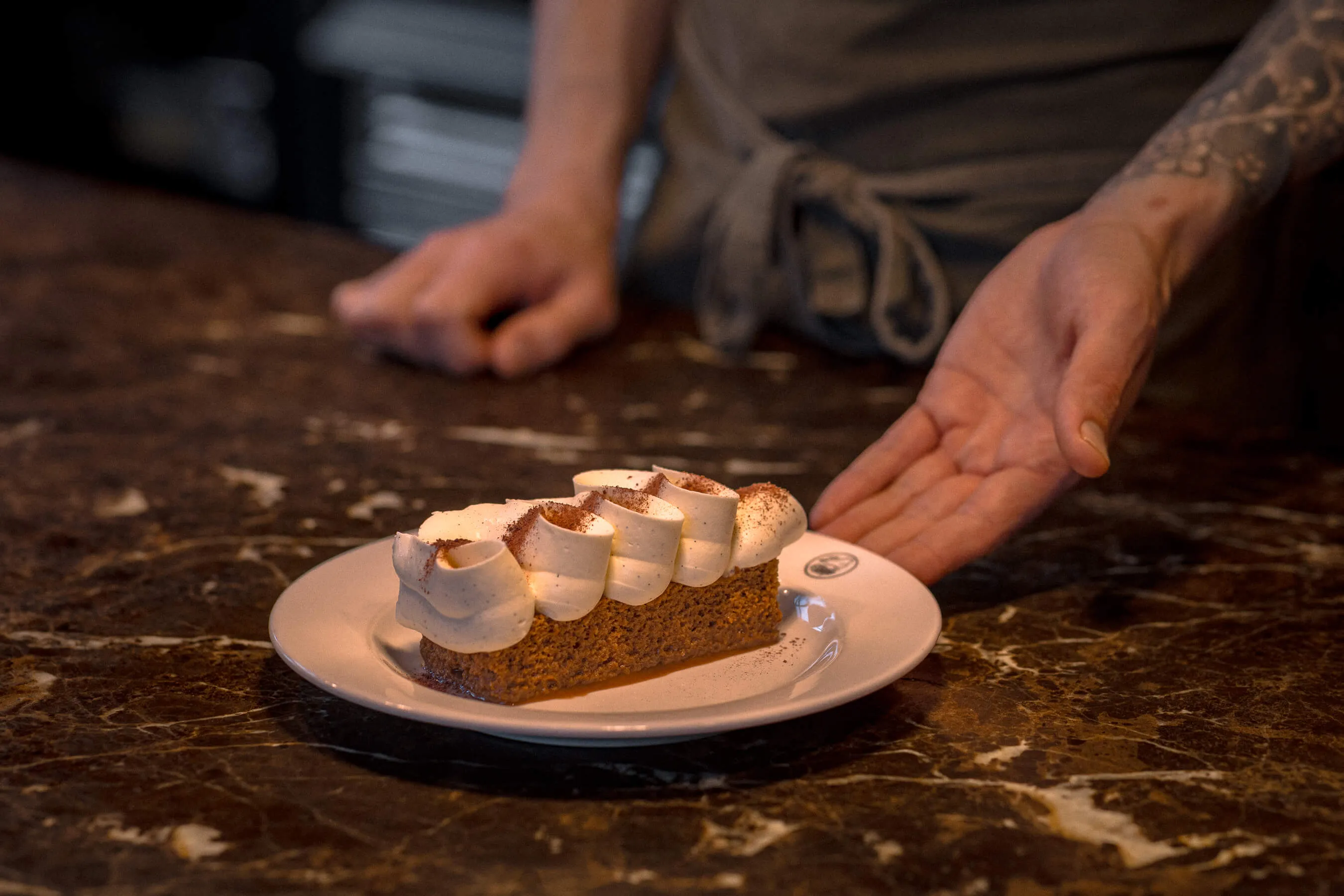 Carrot cake slice topped with rich cream frosting, presented by chef’s tattooed hand at Le Foote on a marble pass | EDG Studios