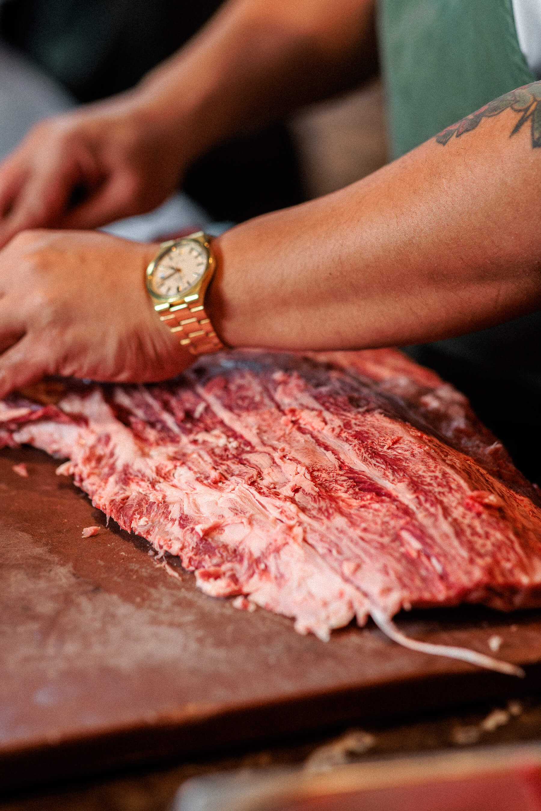 A chef’s tattooed arm and gold wristwatch captured mid-preparation while working with a slab of raw meat in the Le Foote kitchen.