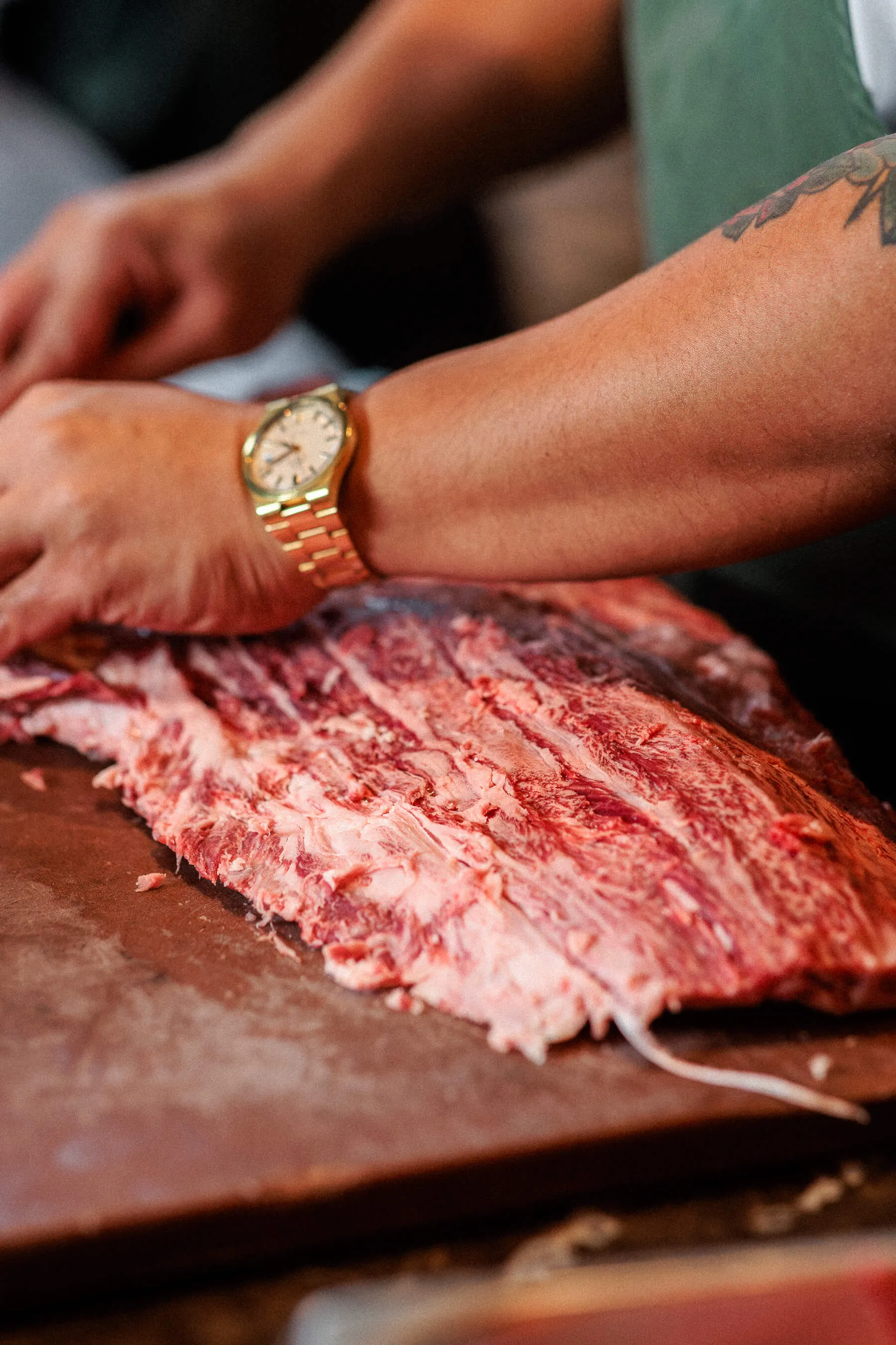 A chef’s tattooed arm and gold wristwatch captured mid-preparation while working with a slab of raw meat in the Le Foote kitchen | EDG Studios