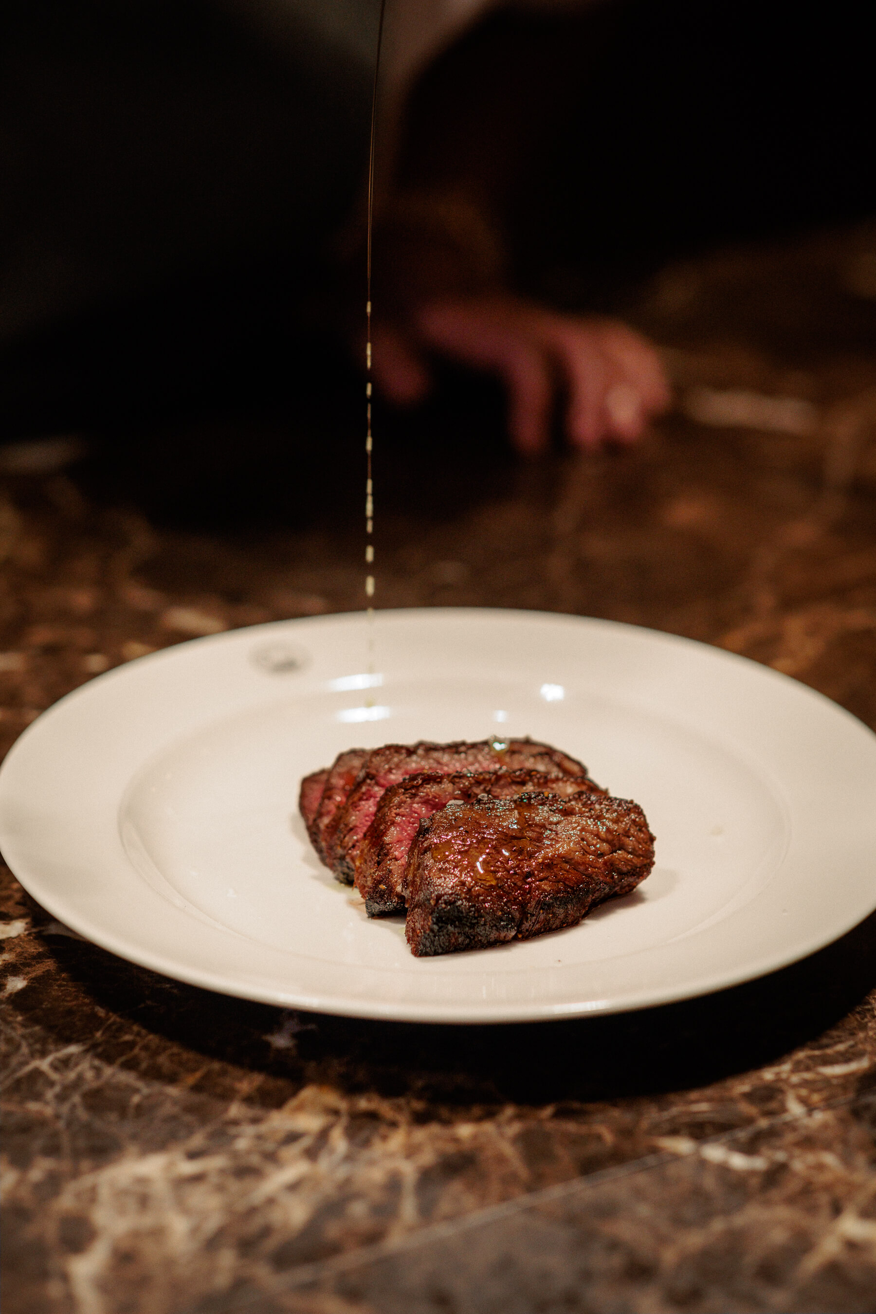 Medium-rare slices of steak plated on a white dish, with lemon juice delicately drizzling from above, capturing a moment of finishing touch.