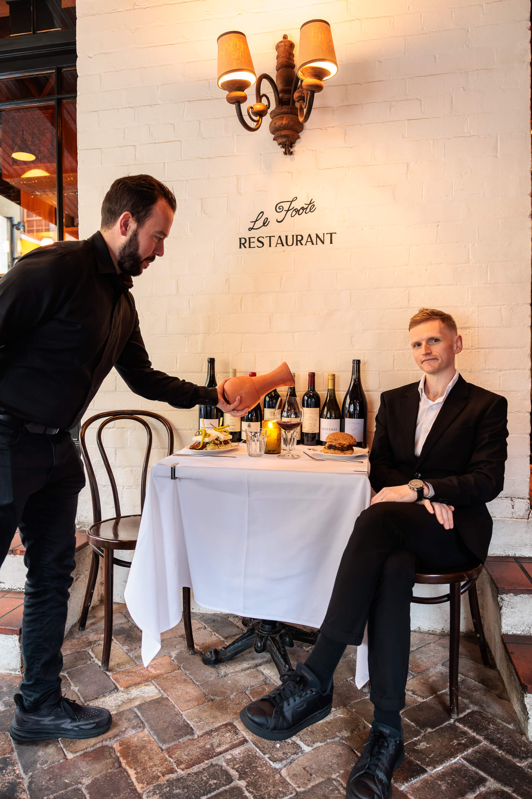 A waiter pours wine for a suited guest seated at a white-tableclothed table outside Le Foote Restaurant, with bottles of wine and plated dishes in the background.