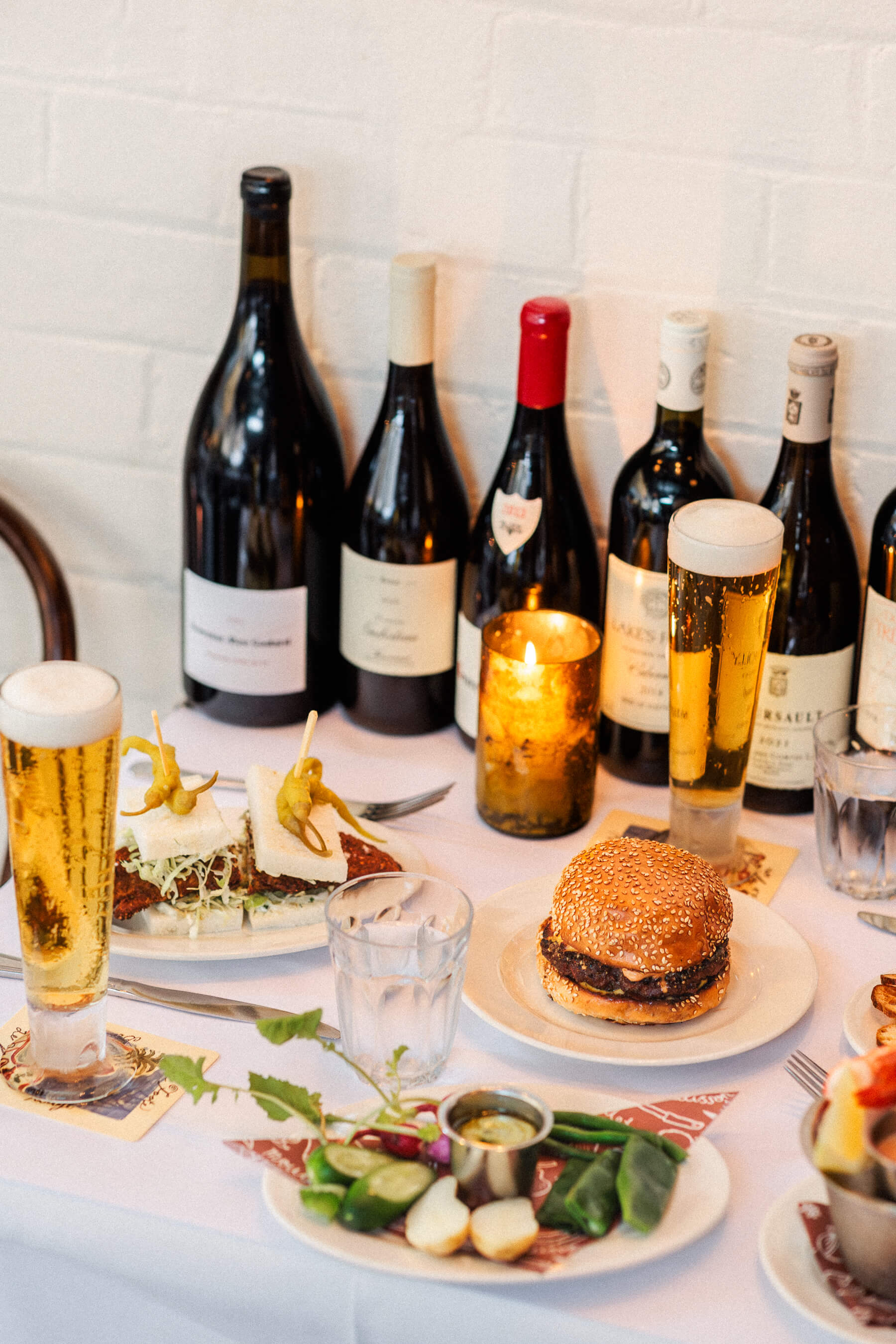 A curated table setting at Le Foote featuring a chicken sandwich, cheeseburger, fresh vegetables, and two tall glasses of beer, backdropped by candlelight and wine bottles.