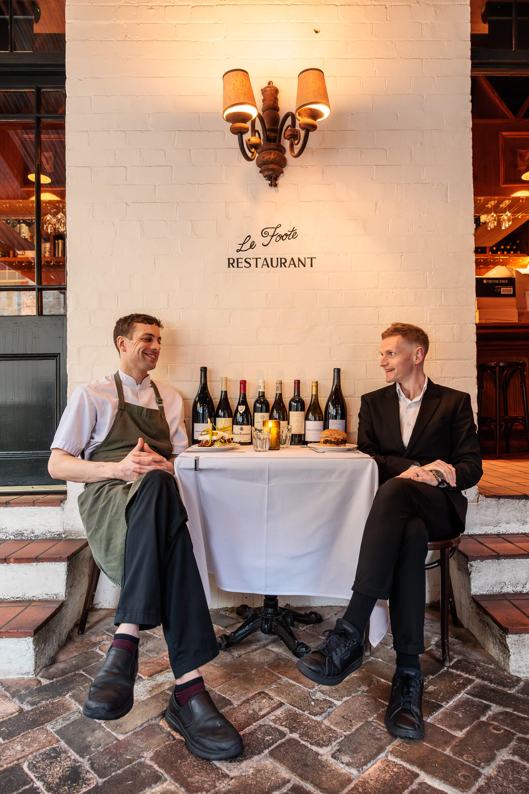 Two men seated at a table outside Le Foote Restaurant in Sydney, sharing a lighthearted moment, surrounded by wine bottles and illuminated by a warm wall sconce.