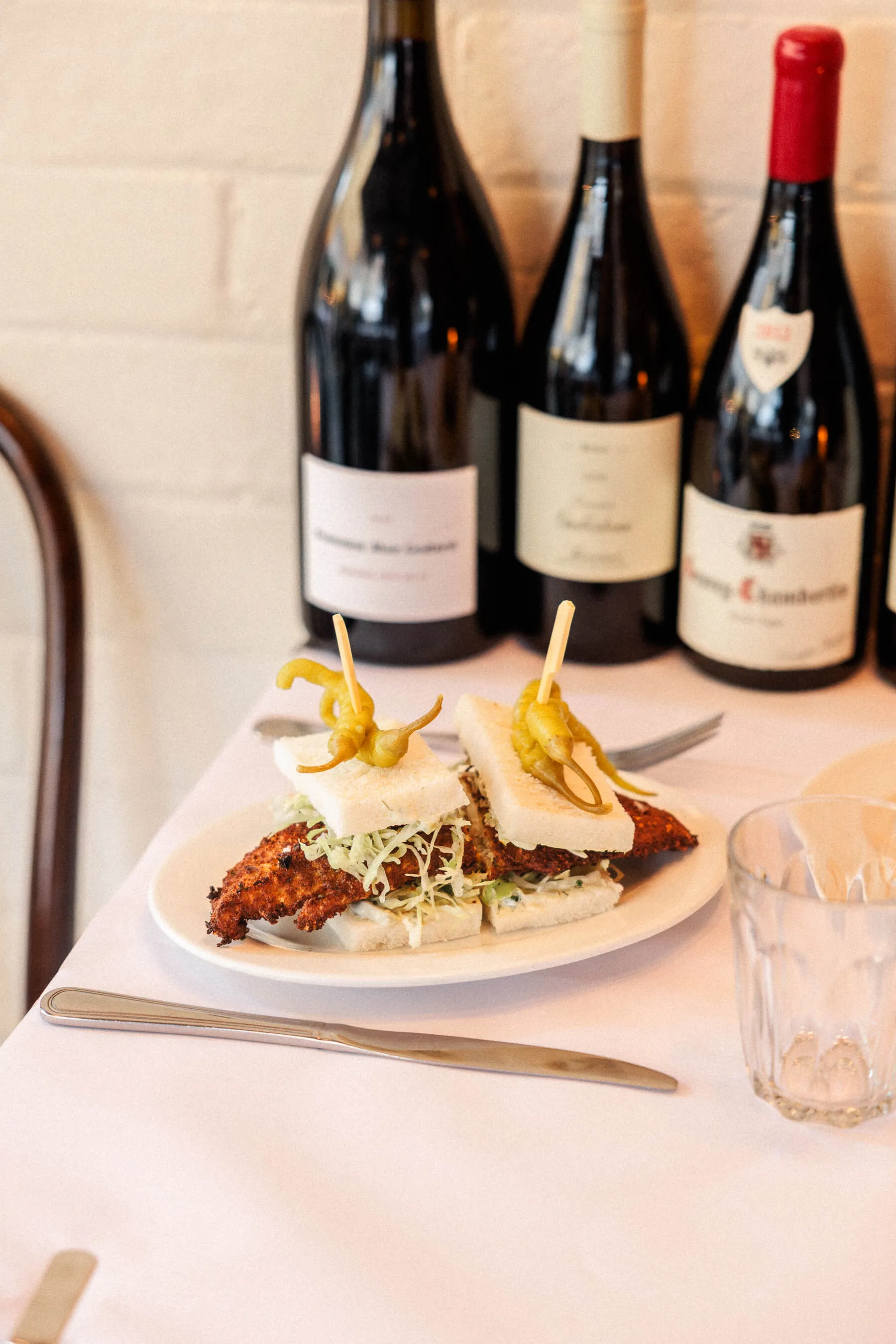 Close-up of a crispy fish sandwich on white bread garnished with pickled peppers, with wine bottles slightly blurred in the background | EDG Studios