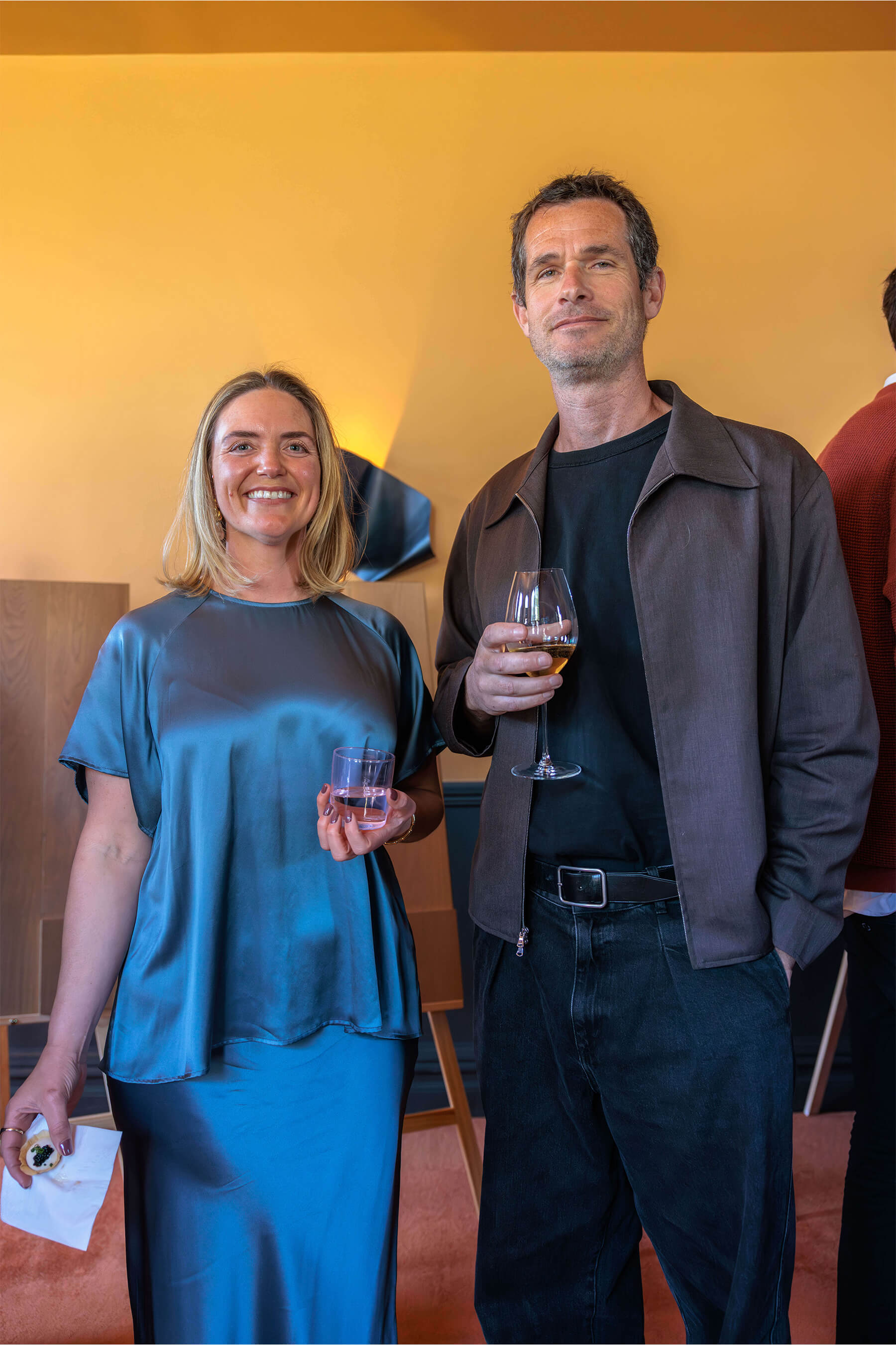 A man and woman standing together at the event, both holding drinks, with wood panel displays and soft lighting in the background.