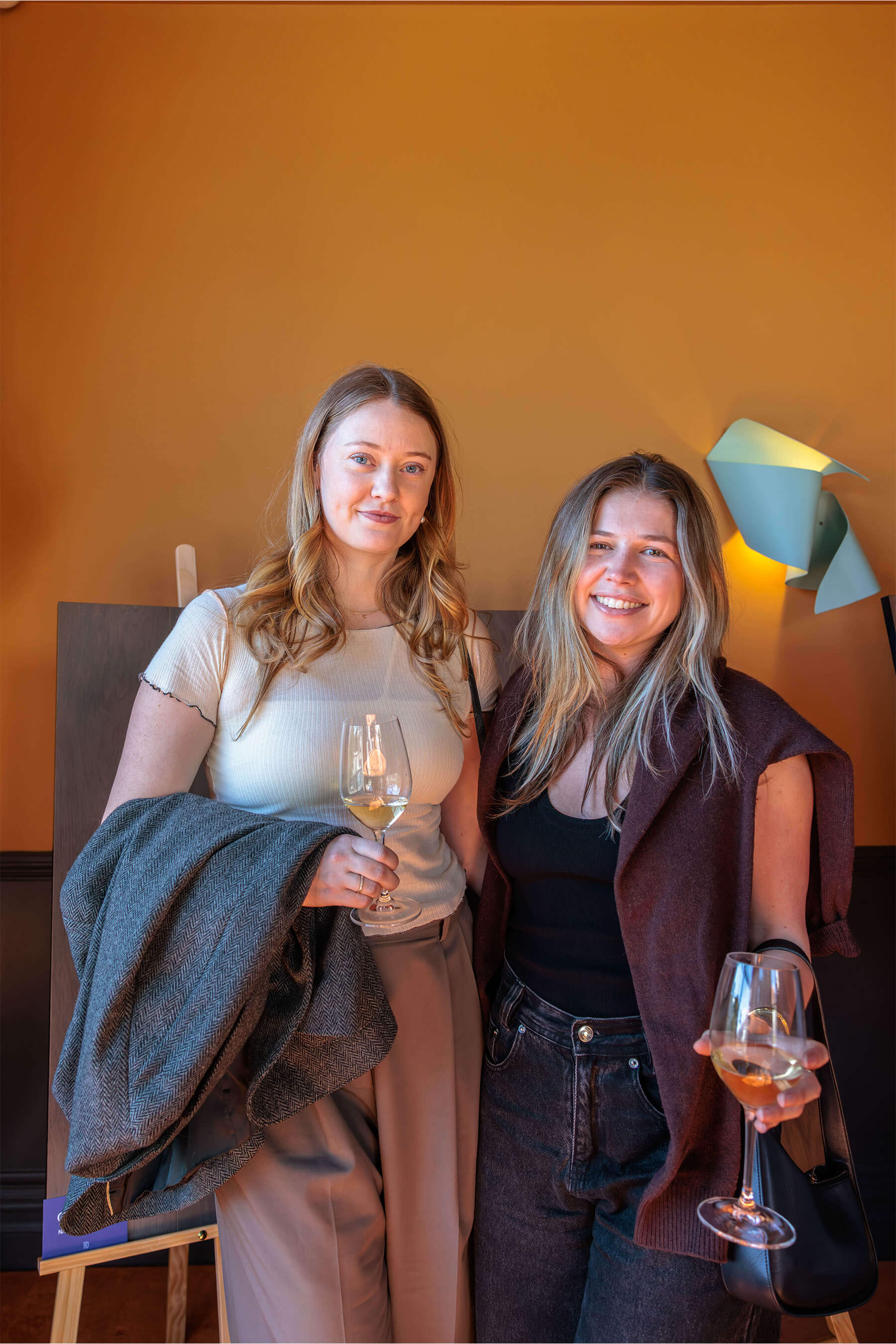 Two women posing with wine glasses in hand, standing in front of sculptural wall lighting and wooden flooring samples at the Royal Oak Floors launch.