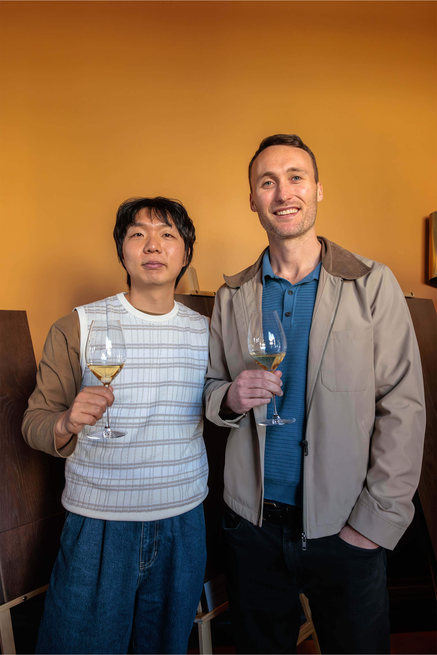 Two men holding wine glasses and smiling at the camera in front of a vibrant orange wall, captured during the Royal Oak Floors product showcase.