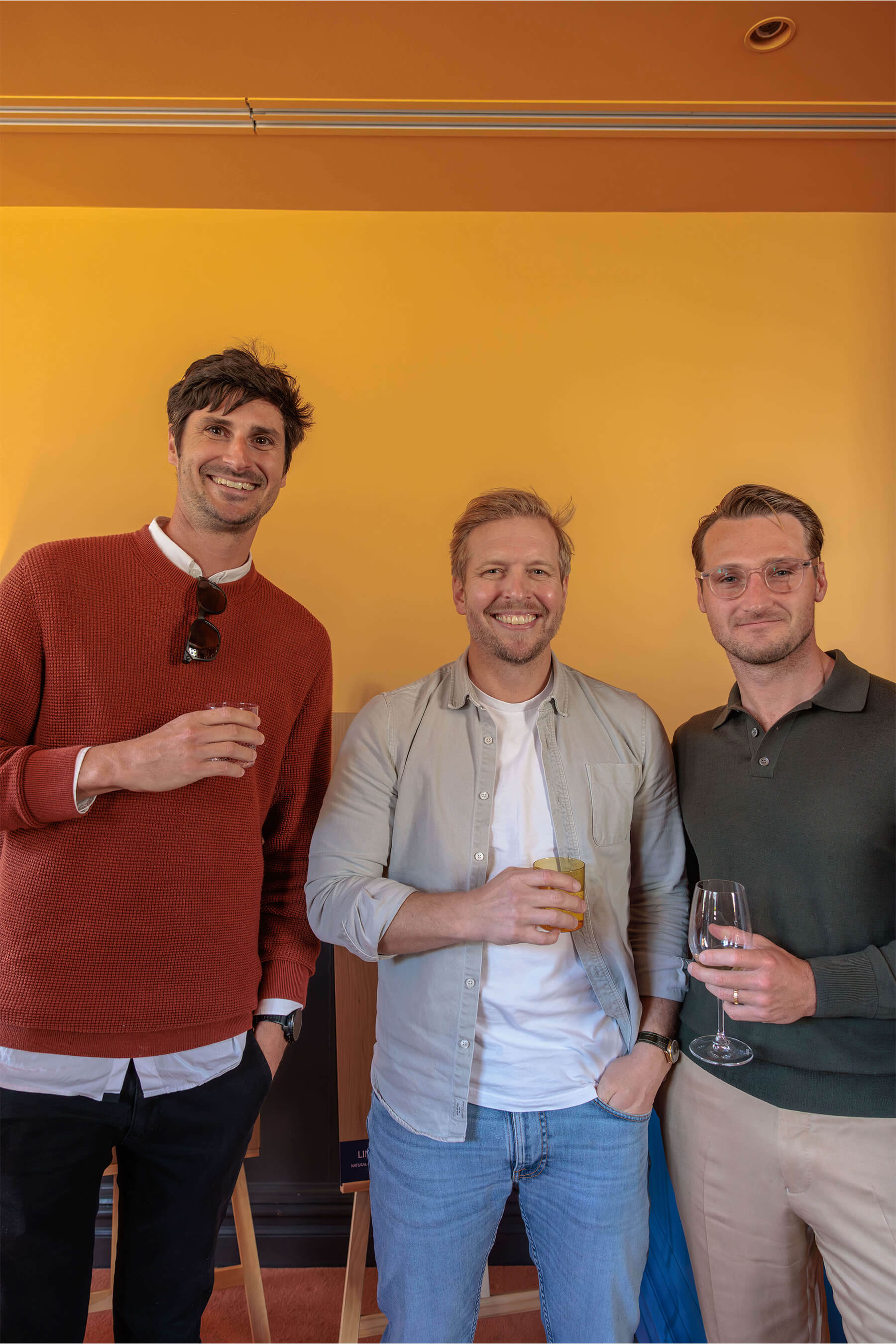 Three men smiling and posing together in front of an ochre wall, each holding a beverage at the Royal Oak Floors launch event in Paddington.