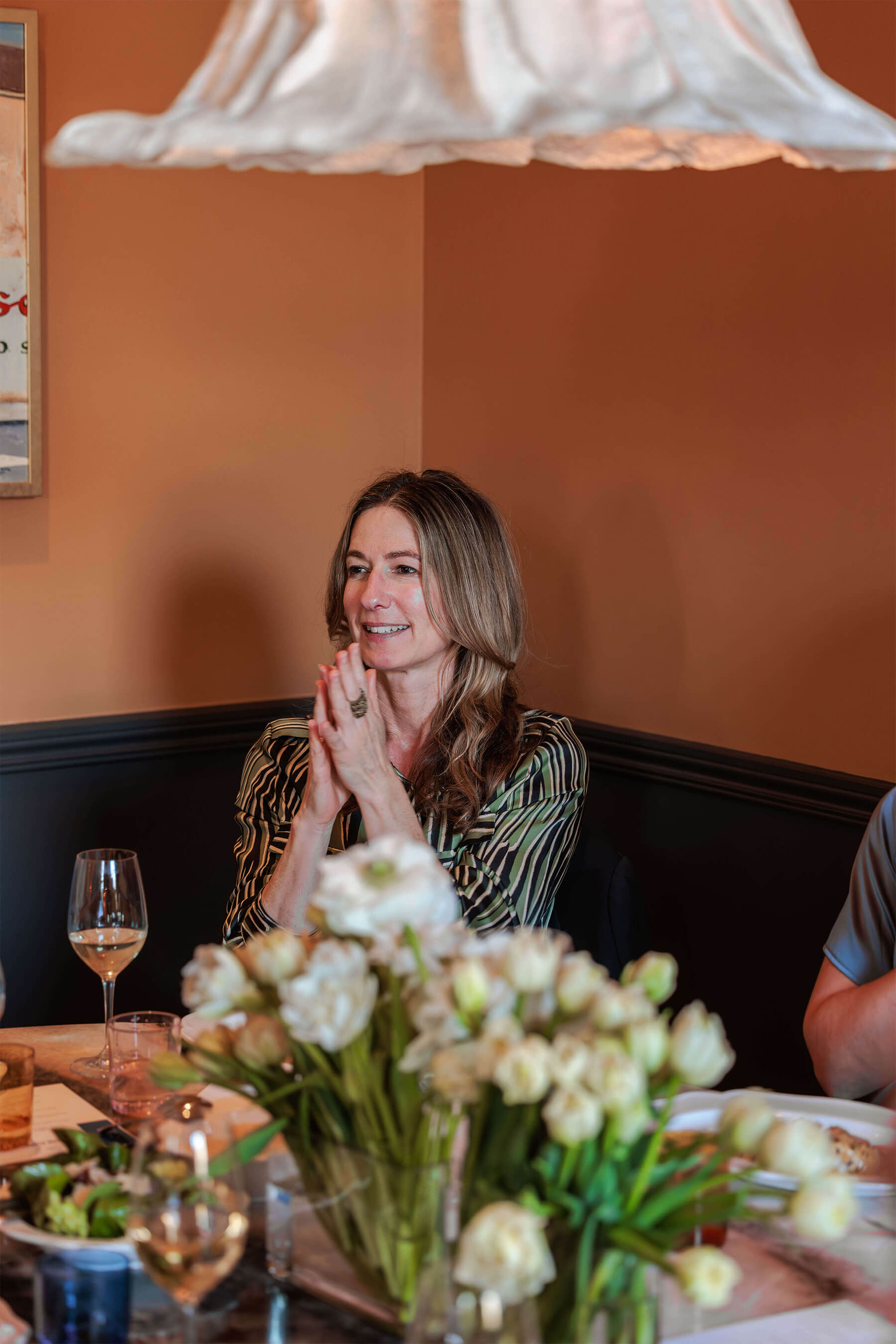 A woman in a green-striped dress claps with appreciation during a speech, seated at a table adorned with fresh white tulips and wine glasses.