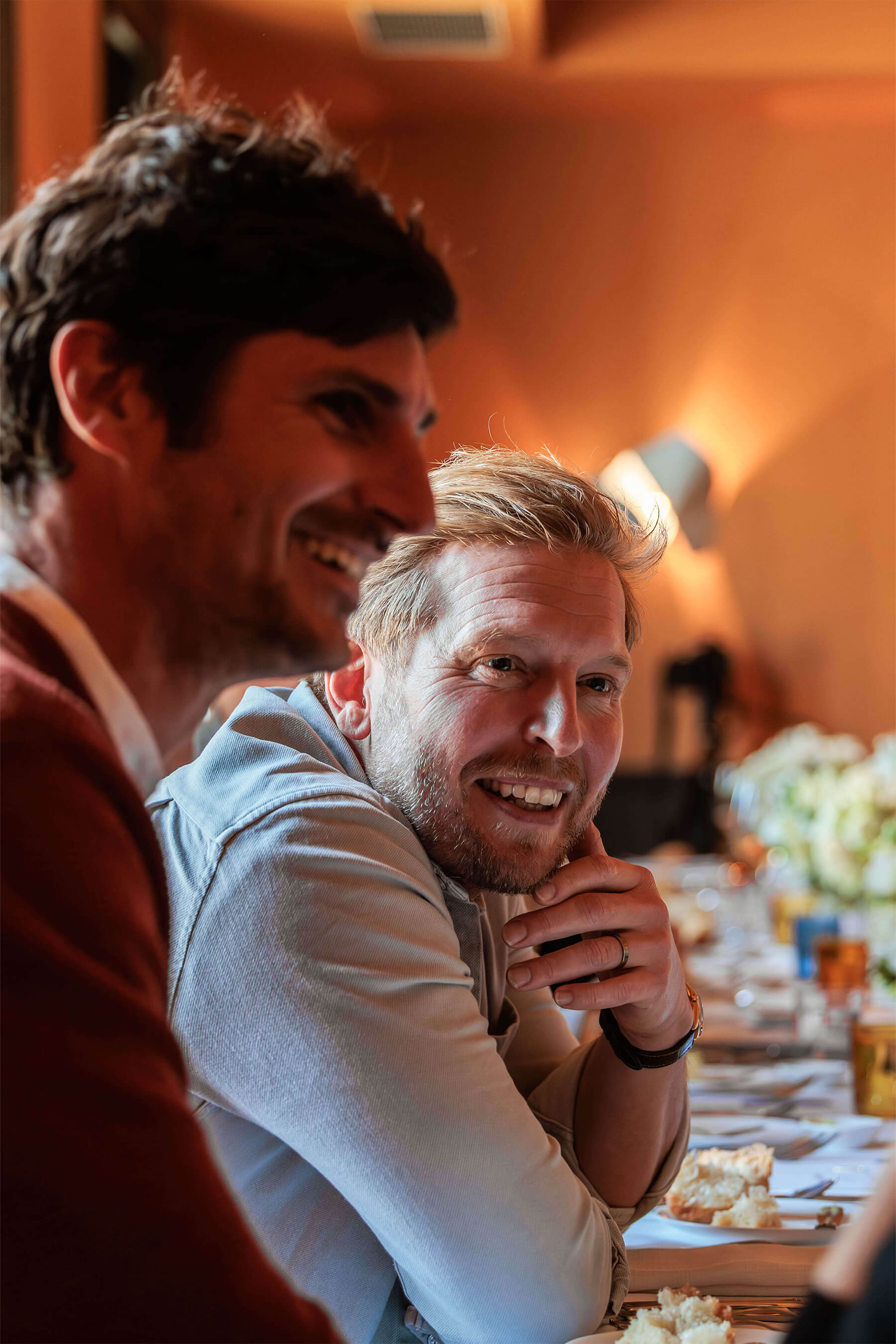 A candid moment of two men laughing and talking during the product launch event, with wine glasses and plates of bread on the table in front of them.