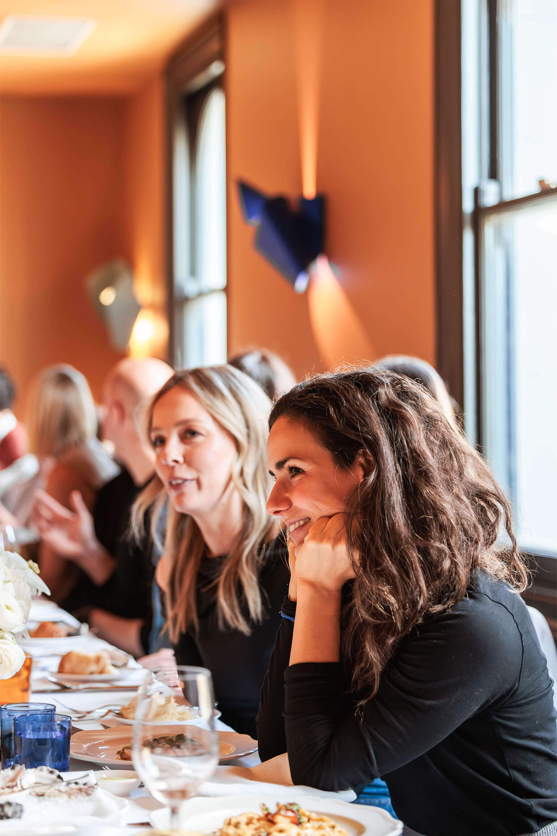 A group of guests dining at the launch event, with a focus on two women in the foreground smiling and chatting at the beautifully set table.