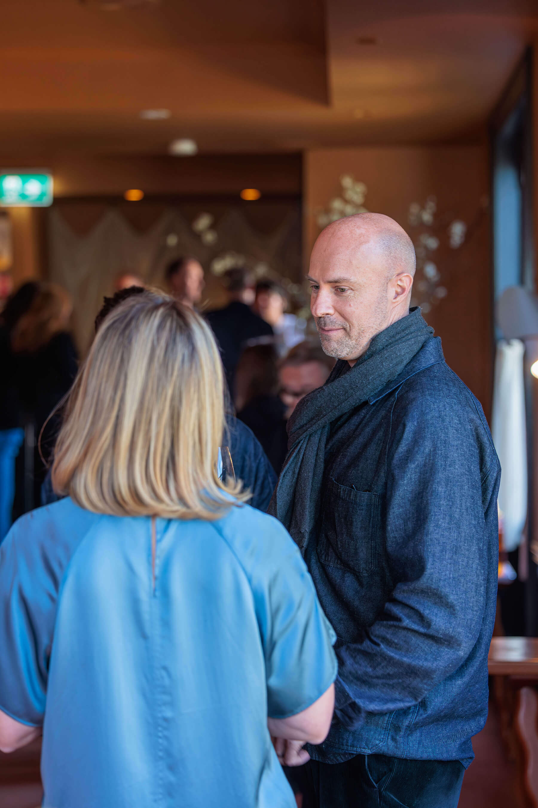 Two guests in conversation during the event, captured from behind one of them in a soft blue blouse, with the crowd blurred in the background.