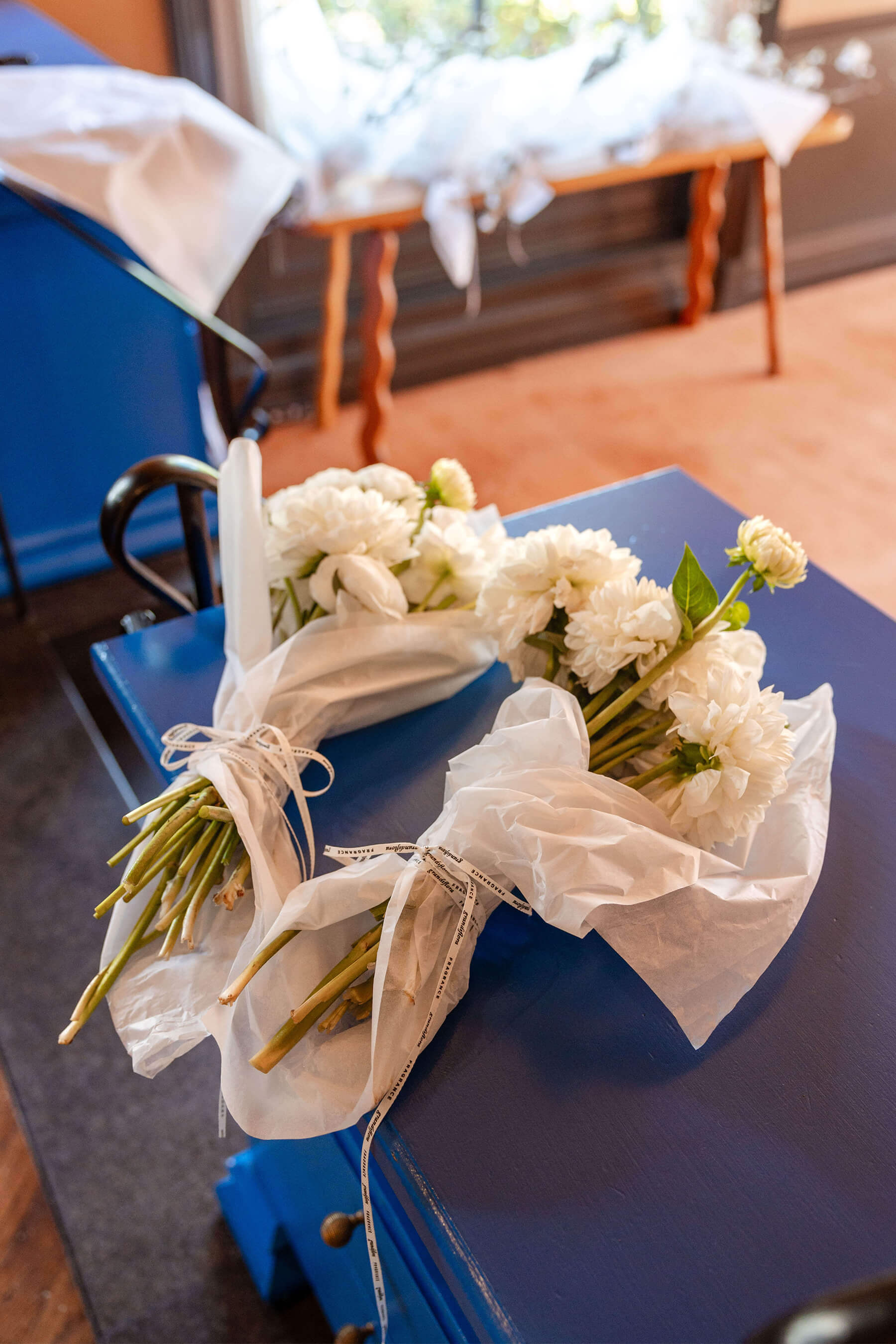 Wrapped floral bouquets with white ranunculus and paper wrapping, laid out on a blue tabletop during event setup.