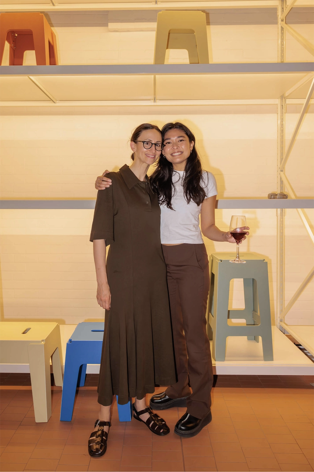 Two attendees smiling together in front of a minimalist shelving display with colourful stools during the DesignByThem speaker series.