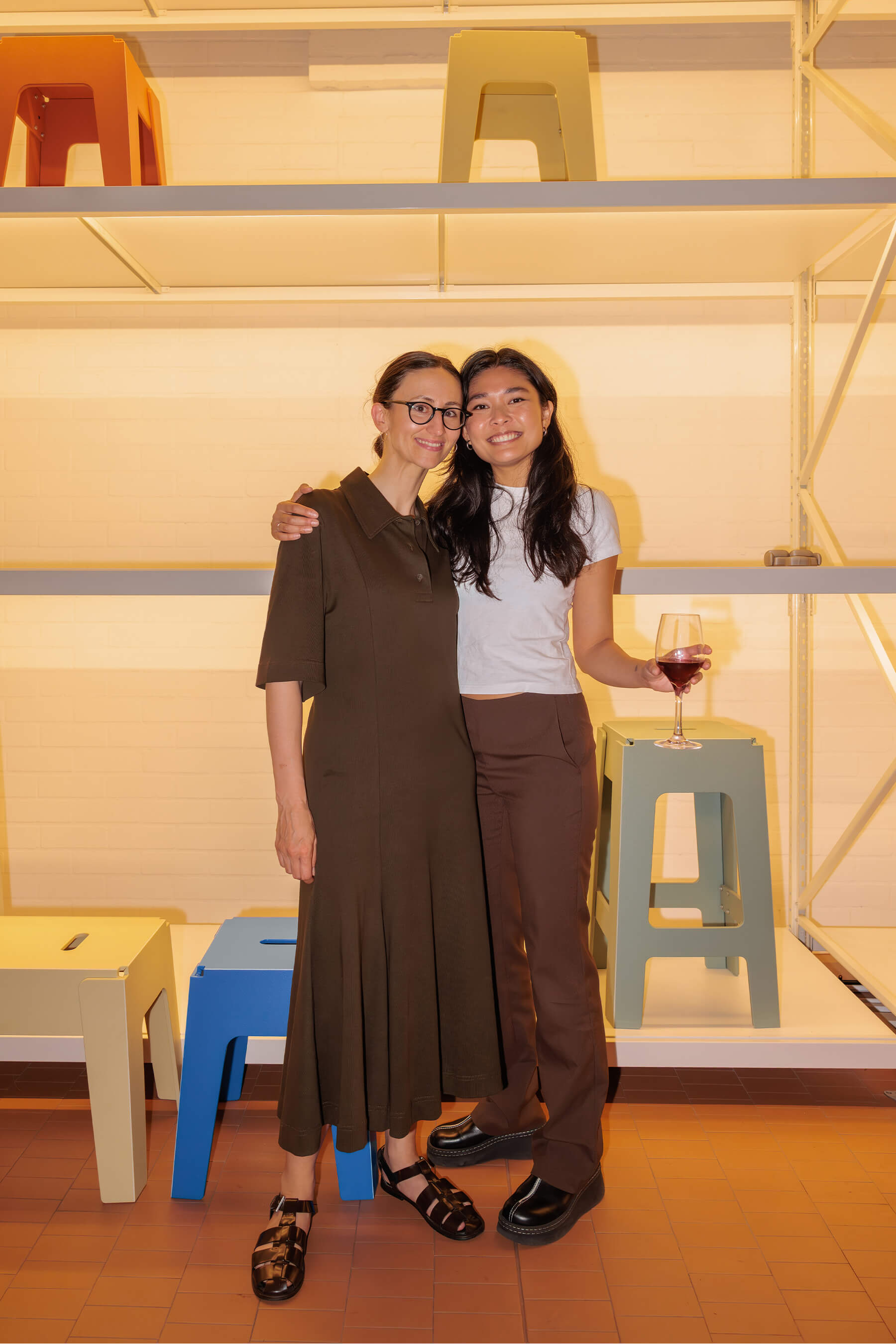 Two attendees smiling together in front of a minimalist shelving display with colourful stools during the DesignByThem speaker series.