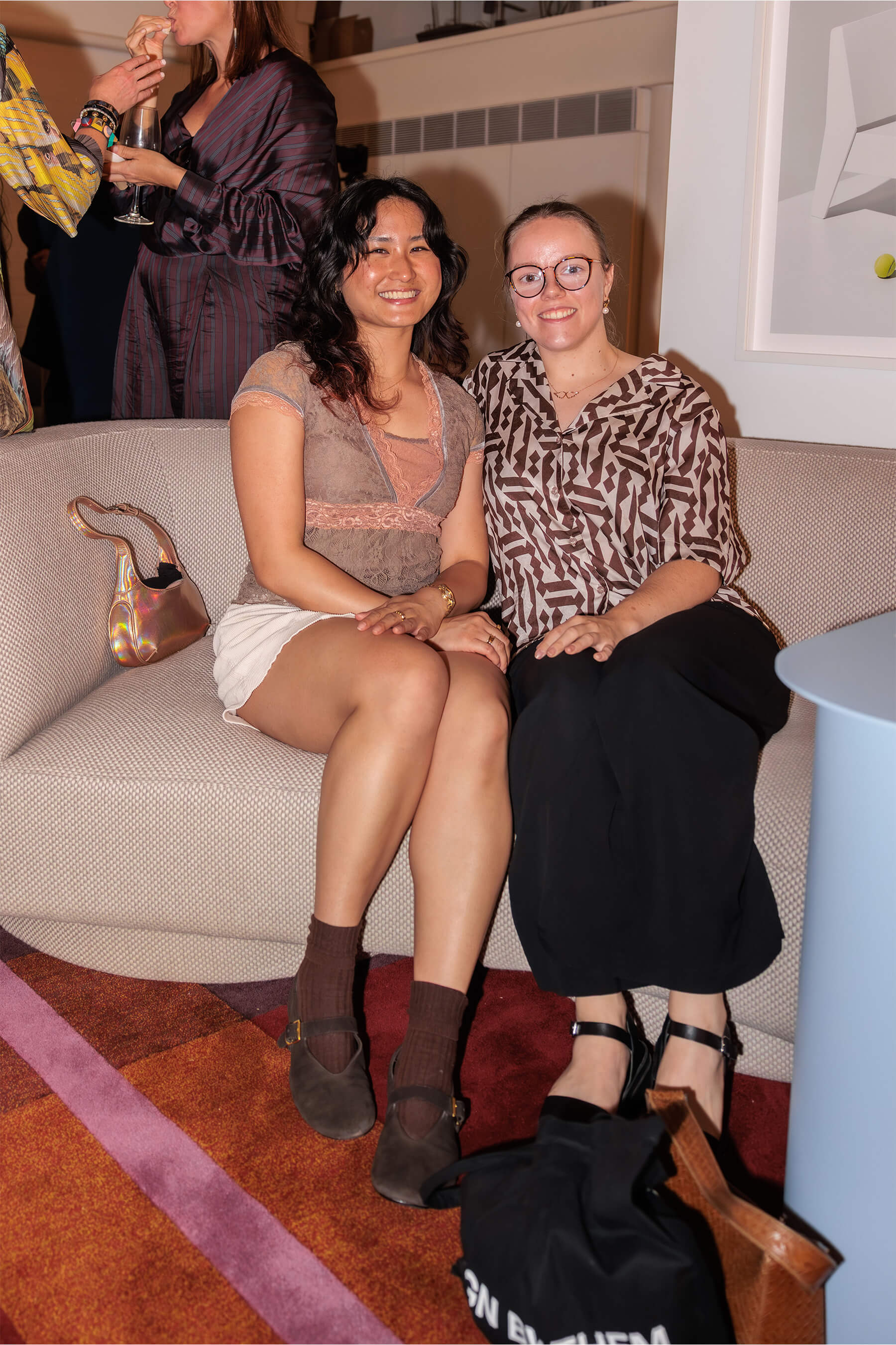 Two guests seated on a beige designer couch at the DesignByThem event, smiling and dressed in relaxed eveningwear.