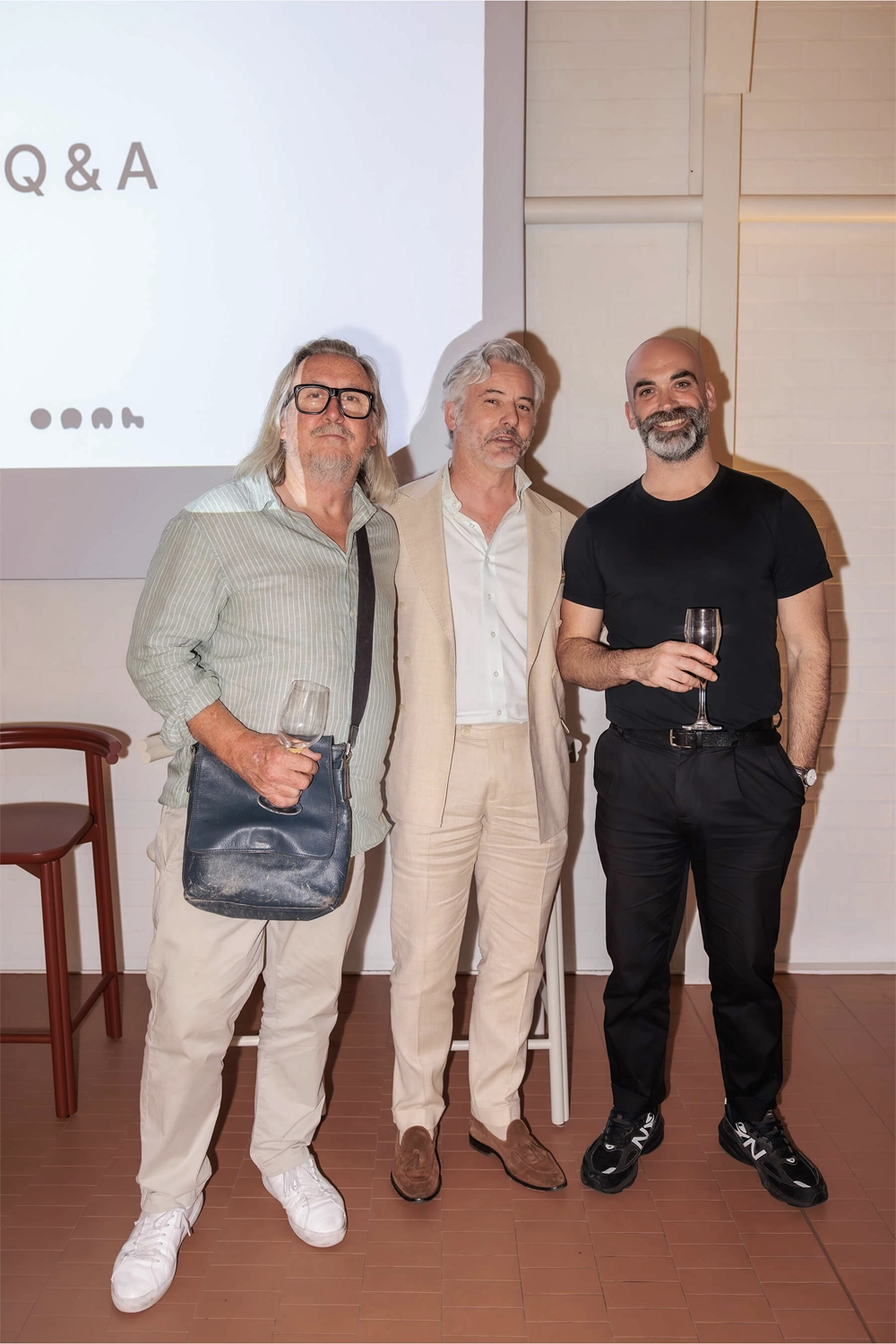 Nicholas Karlovasitis and two other guests pose together in front of a Q&A screen, drinks in hand, at the DesignByThem talk night.