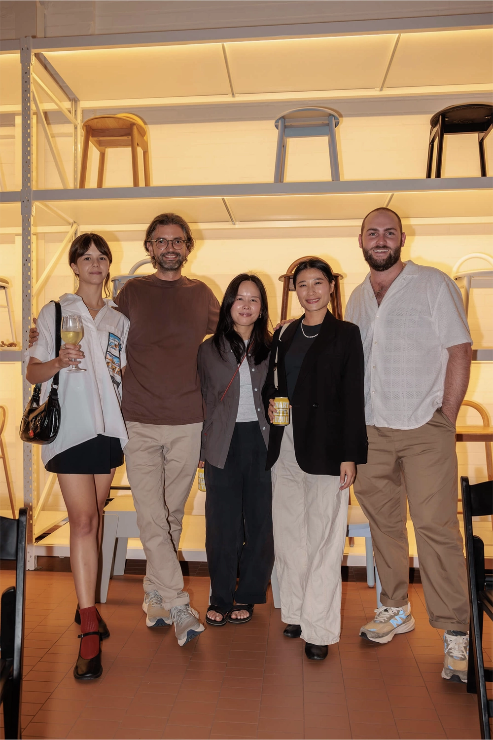 A group of five guests standing together in front of a shelving wall with stools, smiling and enjoying the community spirit of the DesignByThem event.
