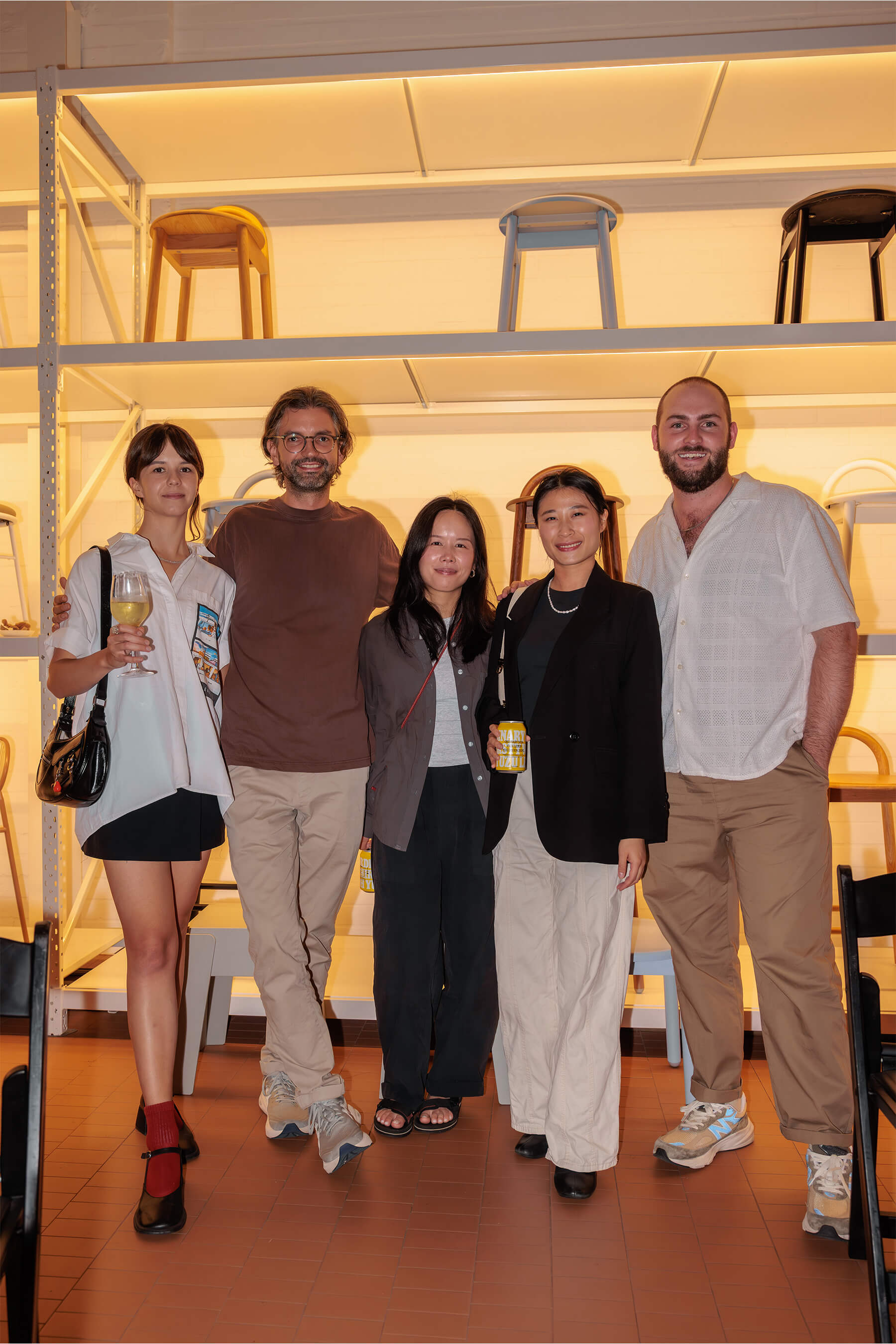A group of five guests standing together in front of a shelving wall with stools, smiling and enjoying the community spirit of the DesignByThem event.