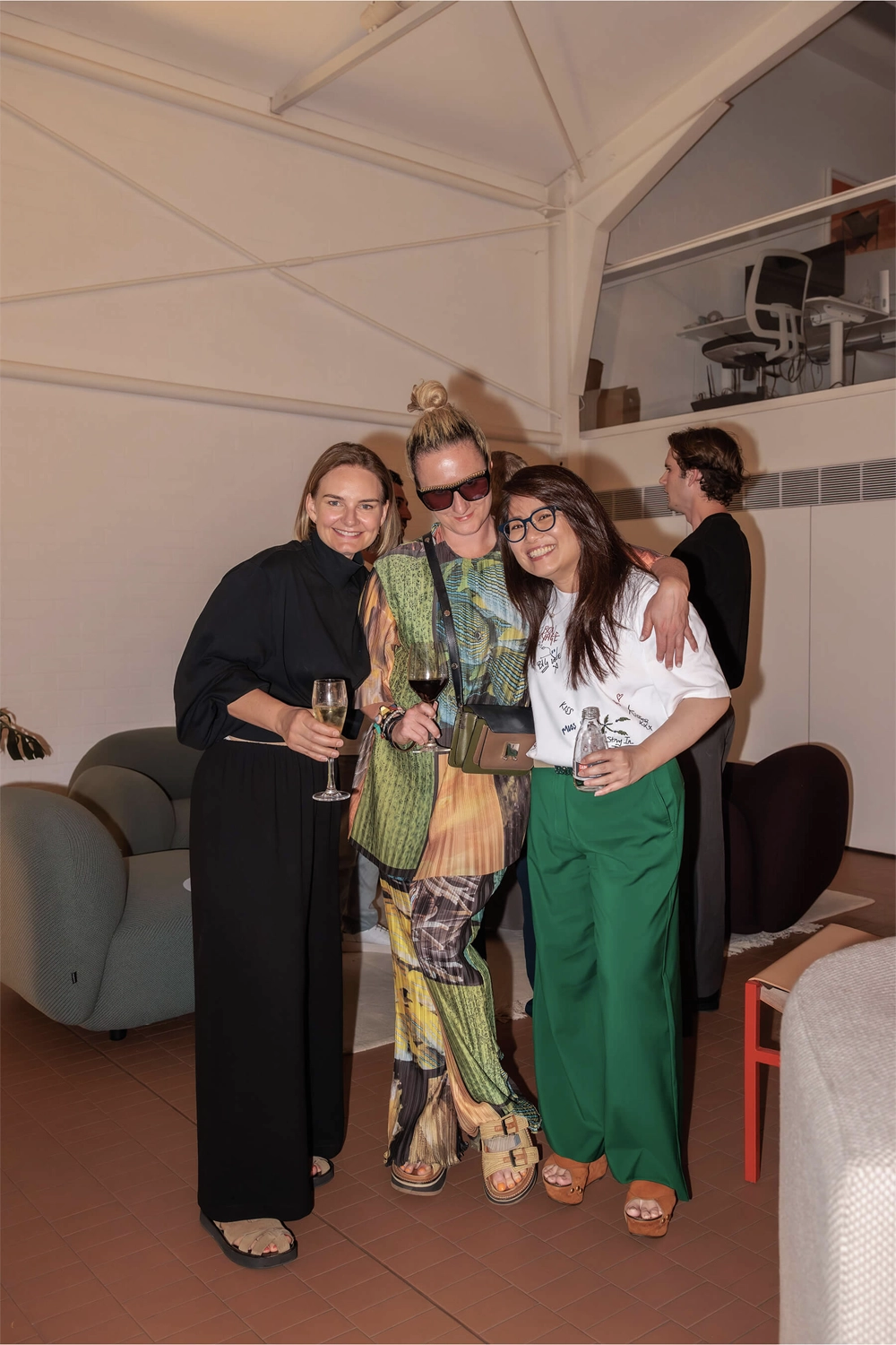 Three women dressed in bold and elegant attire holding wine glasses, laughing and enjoying the DesignByThem event inside a modern showroom.