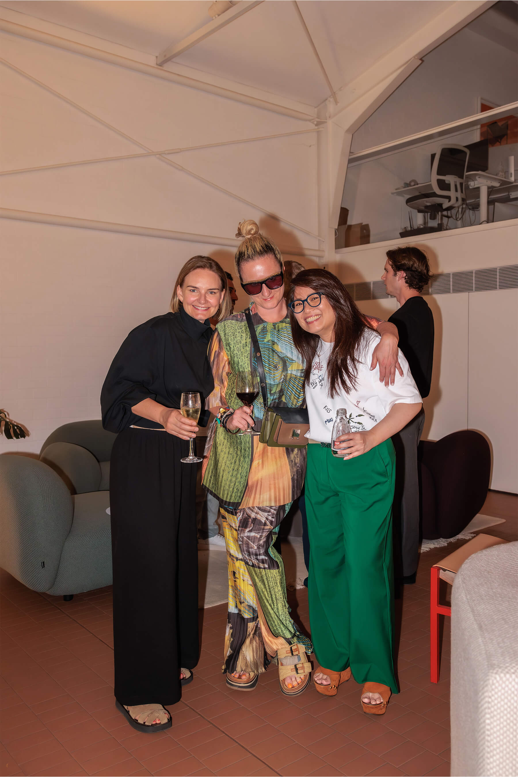 Three women dressed in bold and elegant attire holding wine glasses, laughing and enjoying the DesignByThem event inside a modern showroom.
